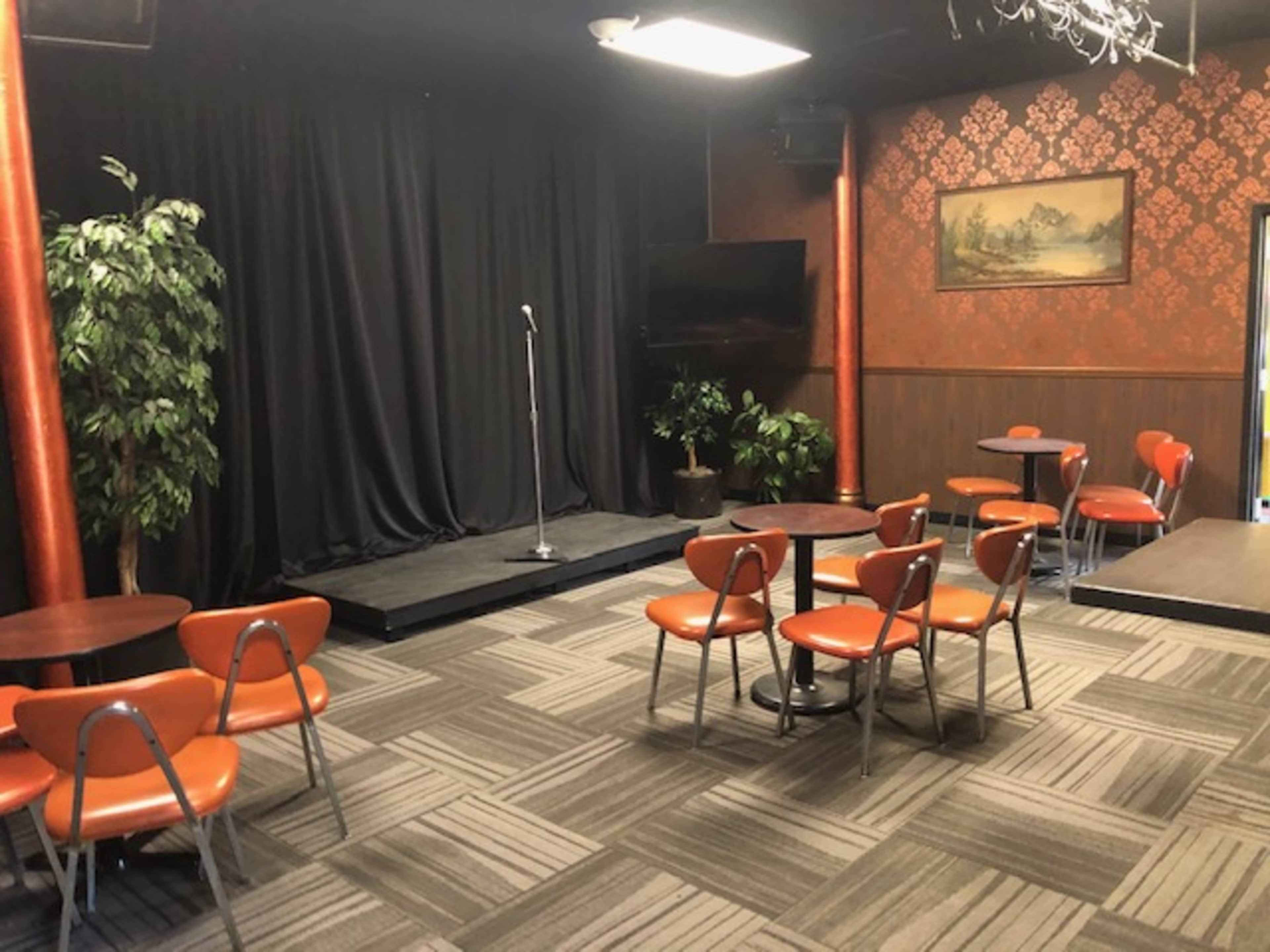 A small performance space with a microphone stand on a raised platform, surrounded by several orange chairs and tables under dim lighting.