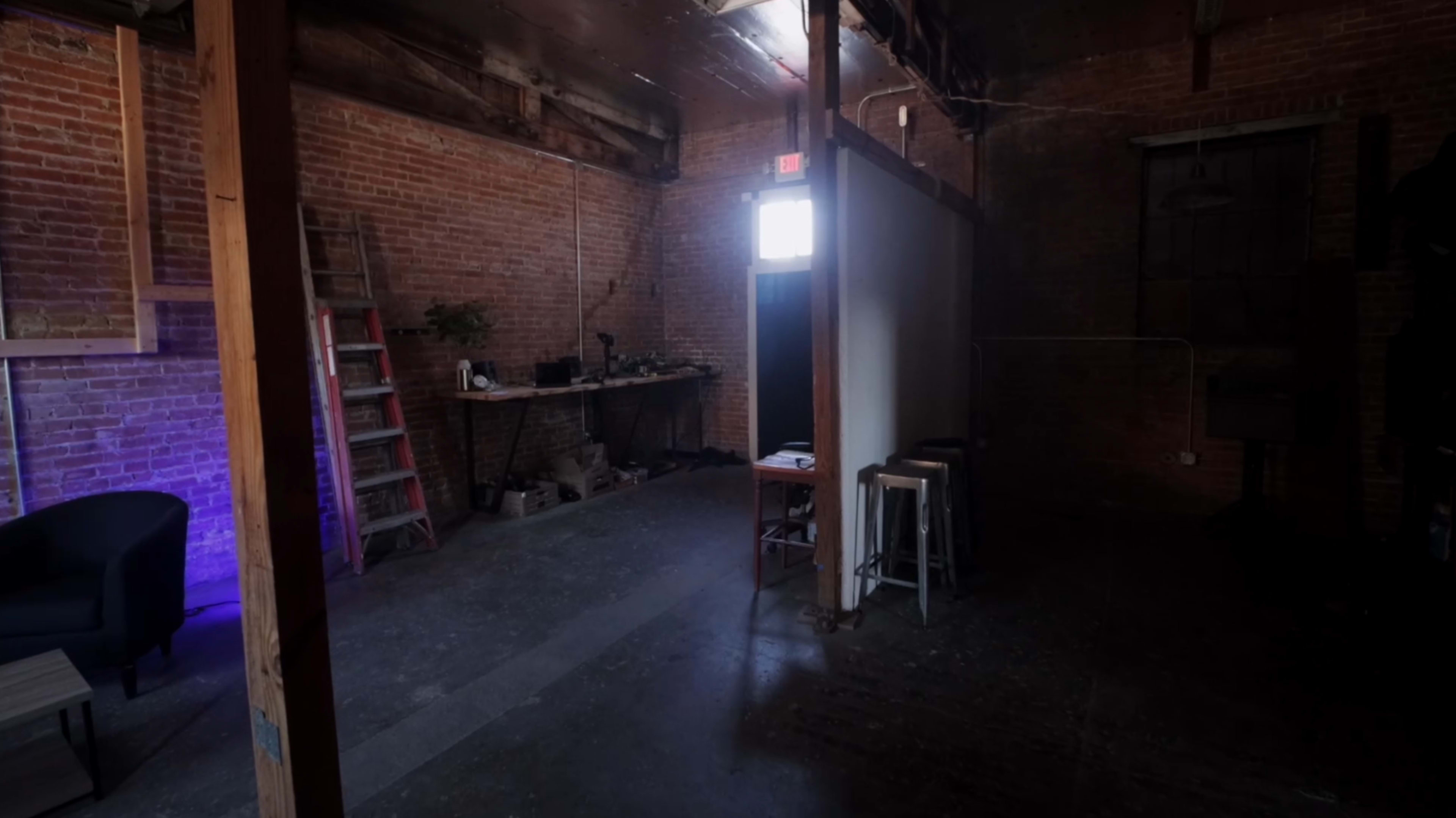 An industrial-style room with brick walls, a ladder, a table with equipment, and a darkened area illuminated by light from an open door.