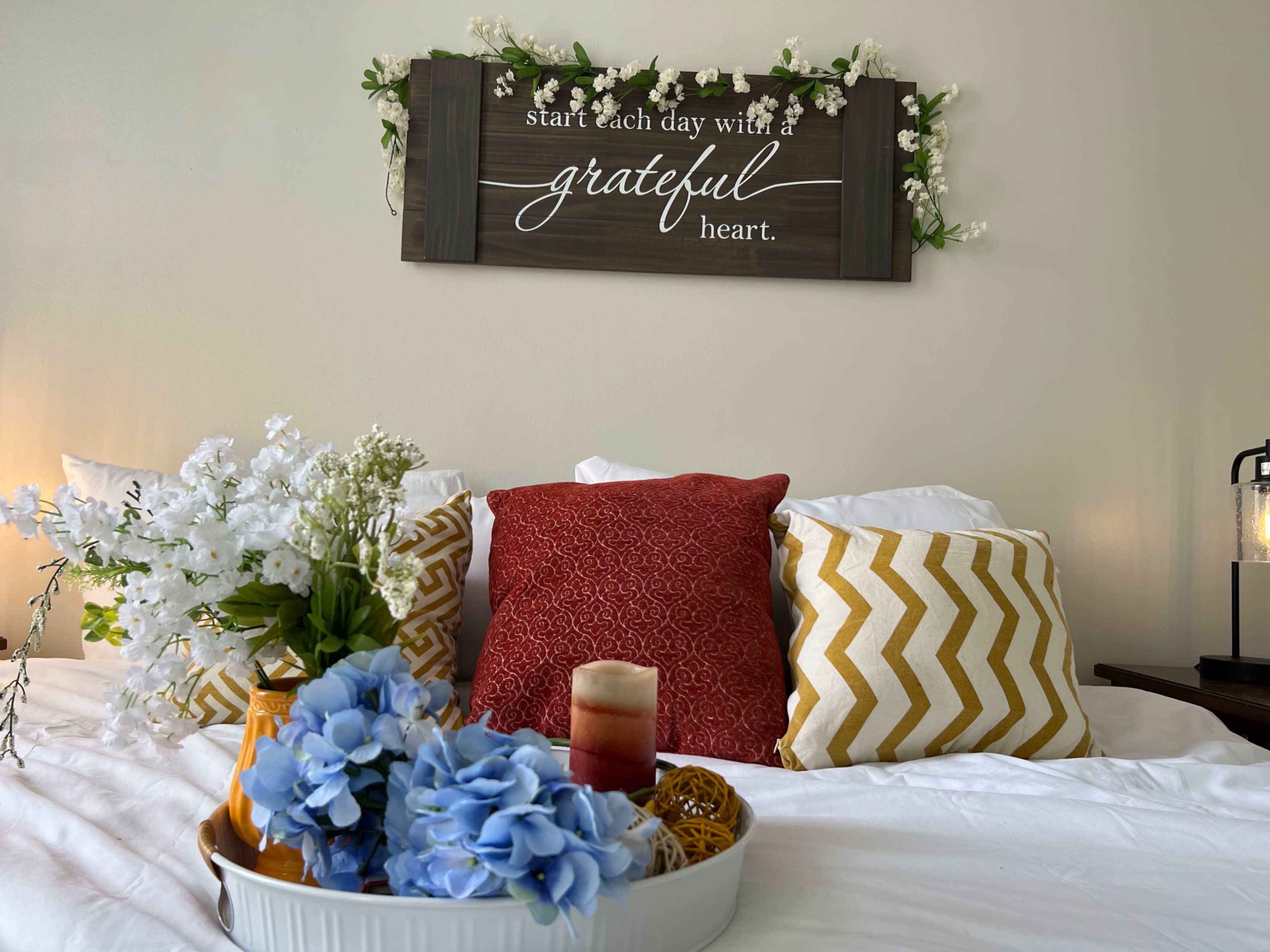 A neatly arranged bed features decorative pillows, a floral centerpiece, and a sign that reads "start each day with a grateful heart."
