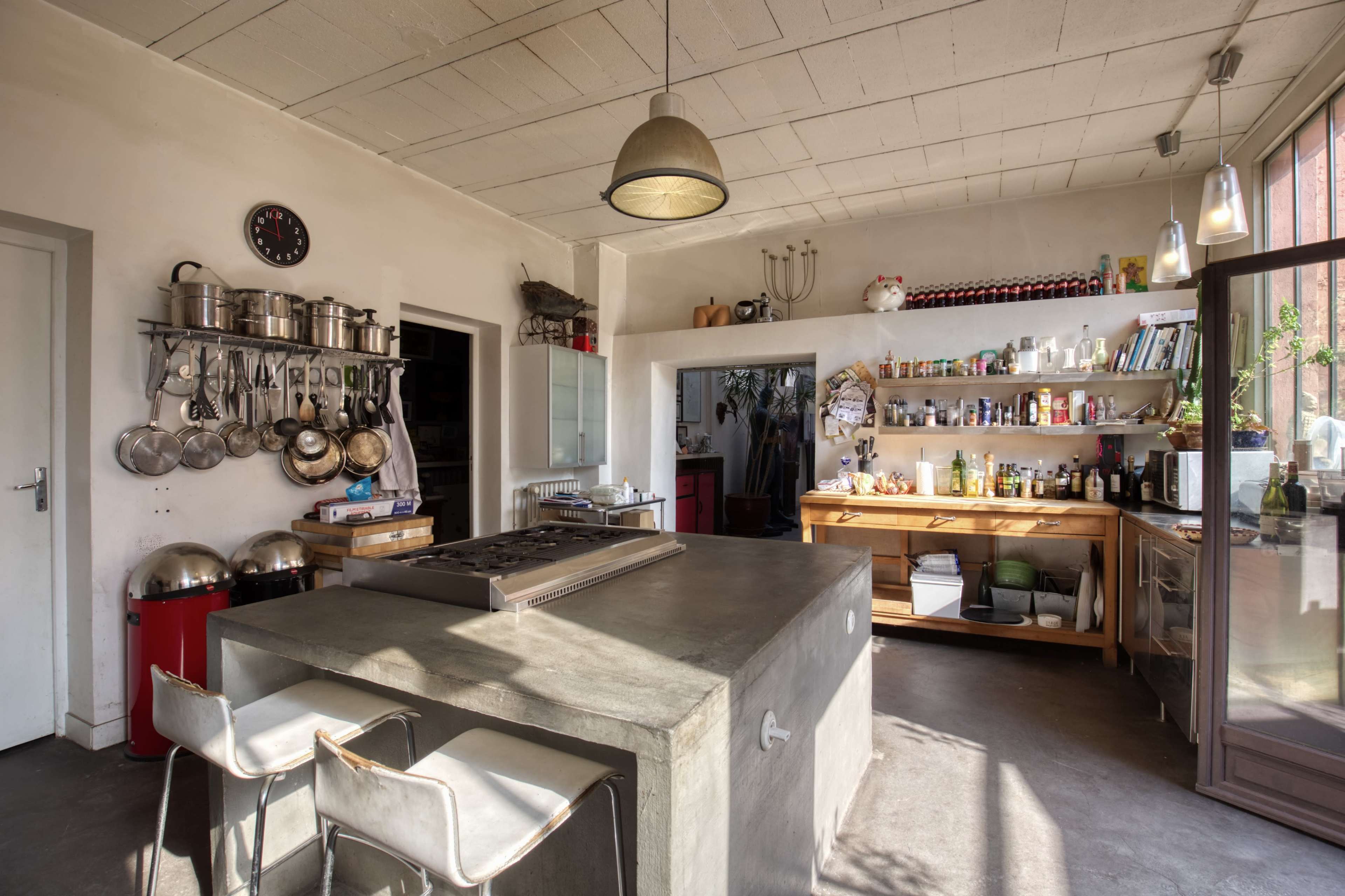 A modern kitchen with a concrete island, stainless steel pots hanging from the wall, and a wooden countertop filled with various cooking ingredients and bottles.