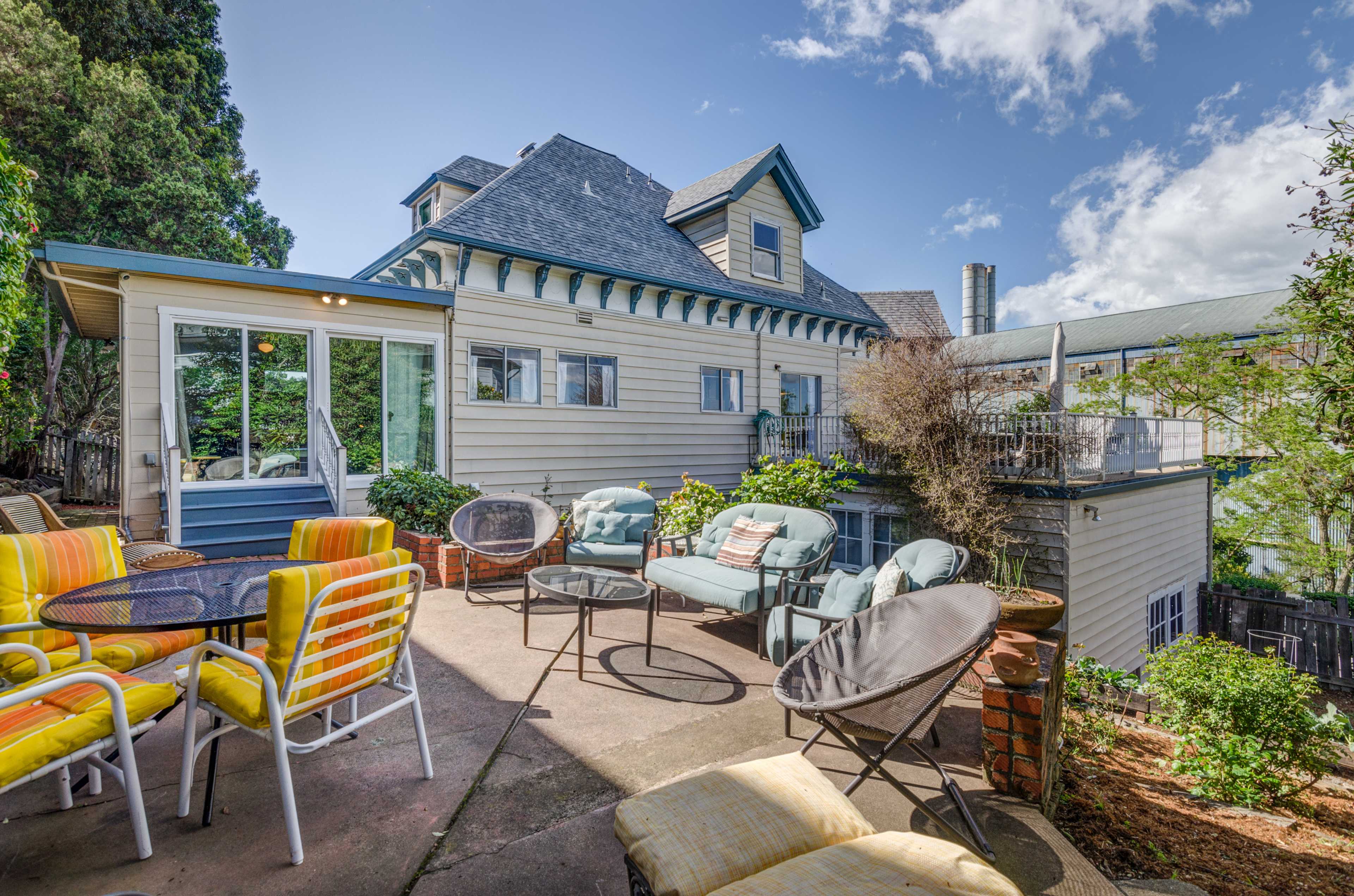 A patio with various seating options is situated behind a multi-story house surrounded by greenery and blue skies.
