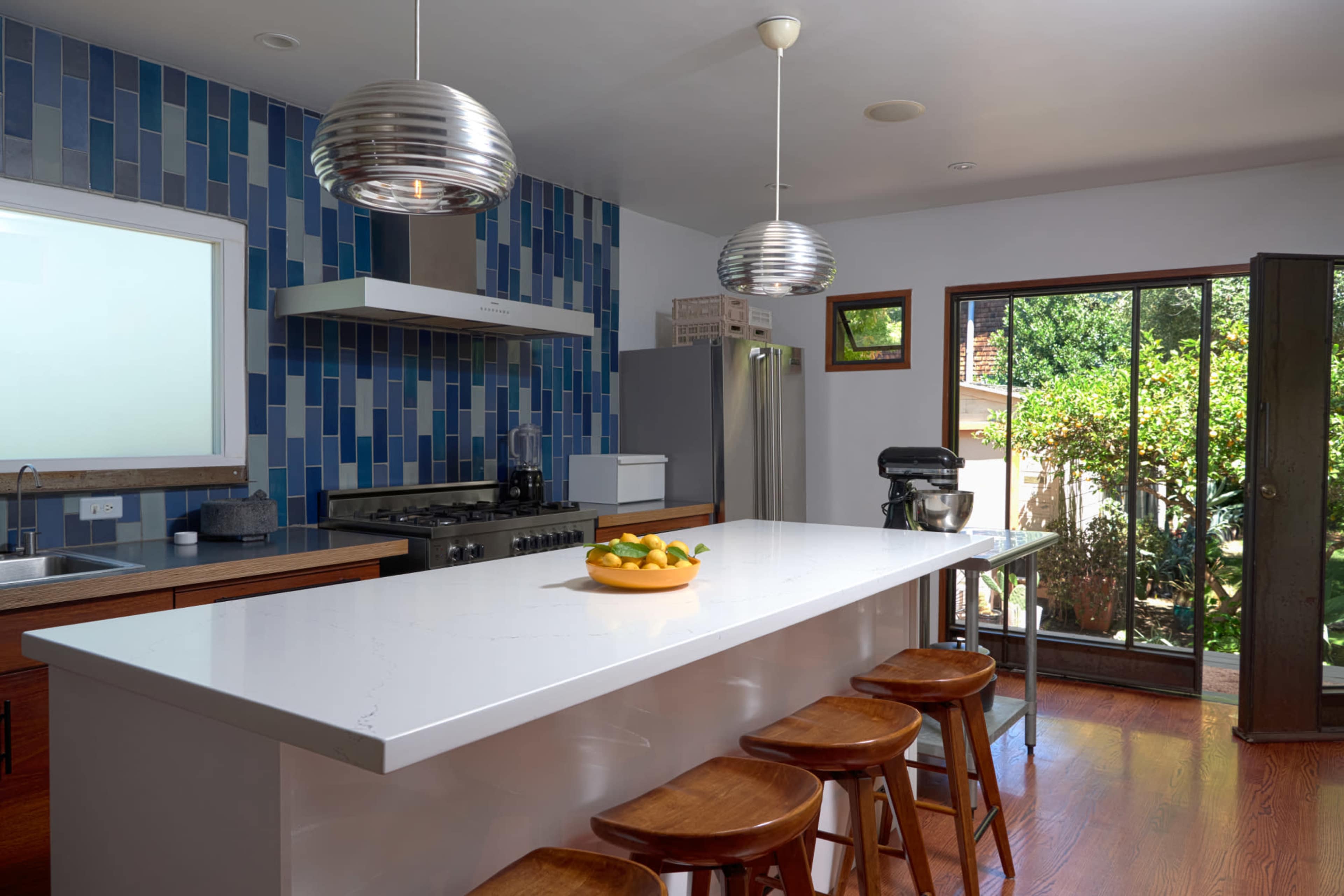 A modern kitchen features a white island with a bowl of fruit, metal pendant lights, and blue tile accents on the walls.