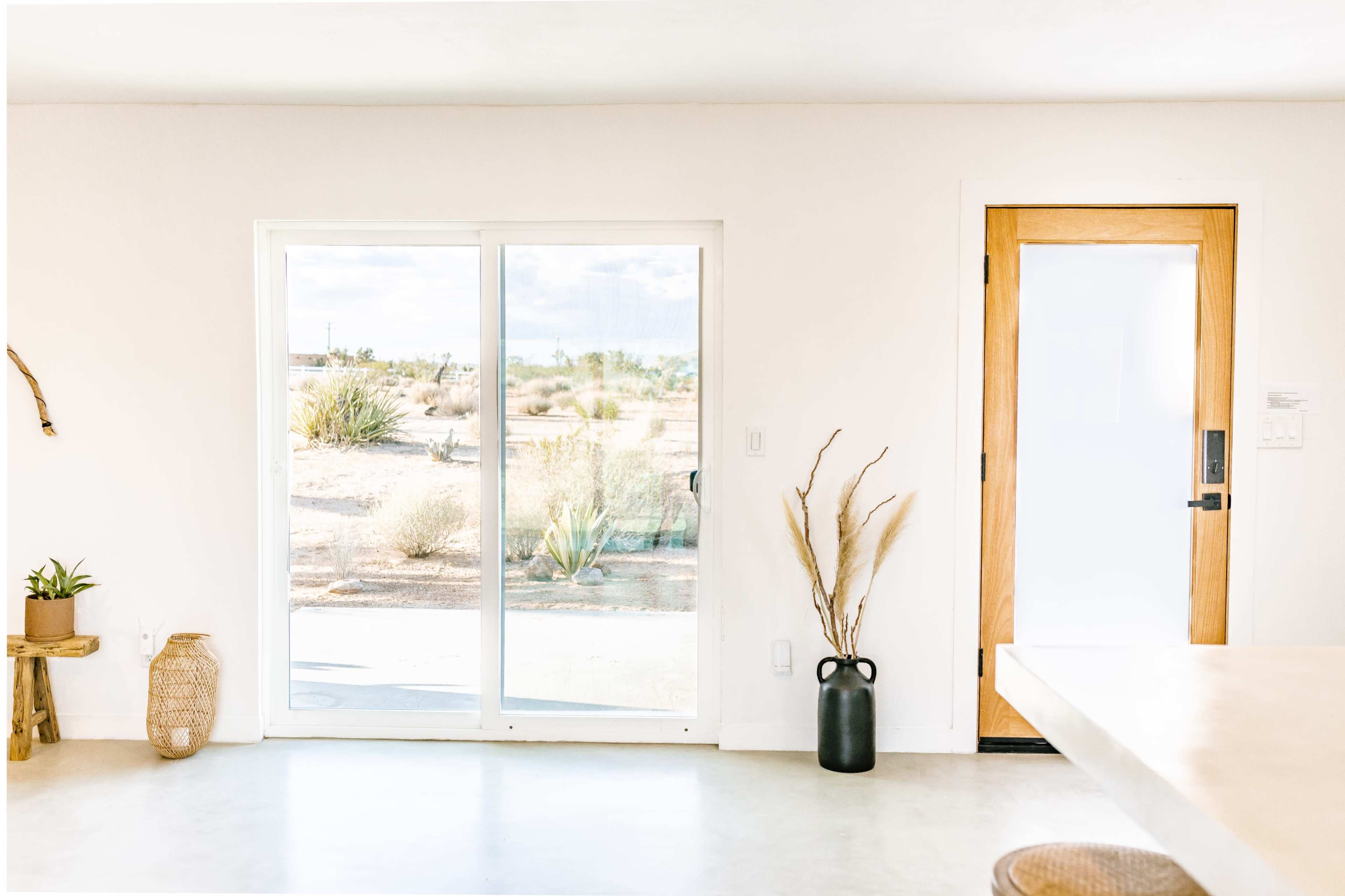 The scene shows a modern interior with a large sliding glass door that opens to a desert landscape, featuring minimal furnishings and natural decor elements.