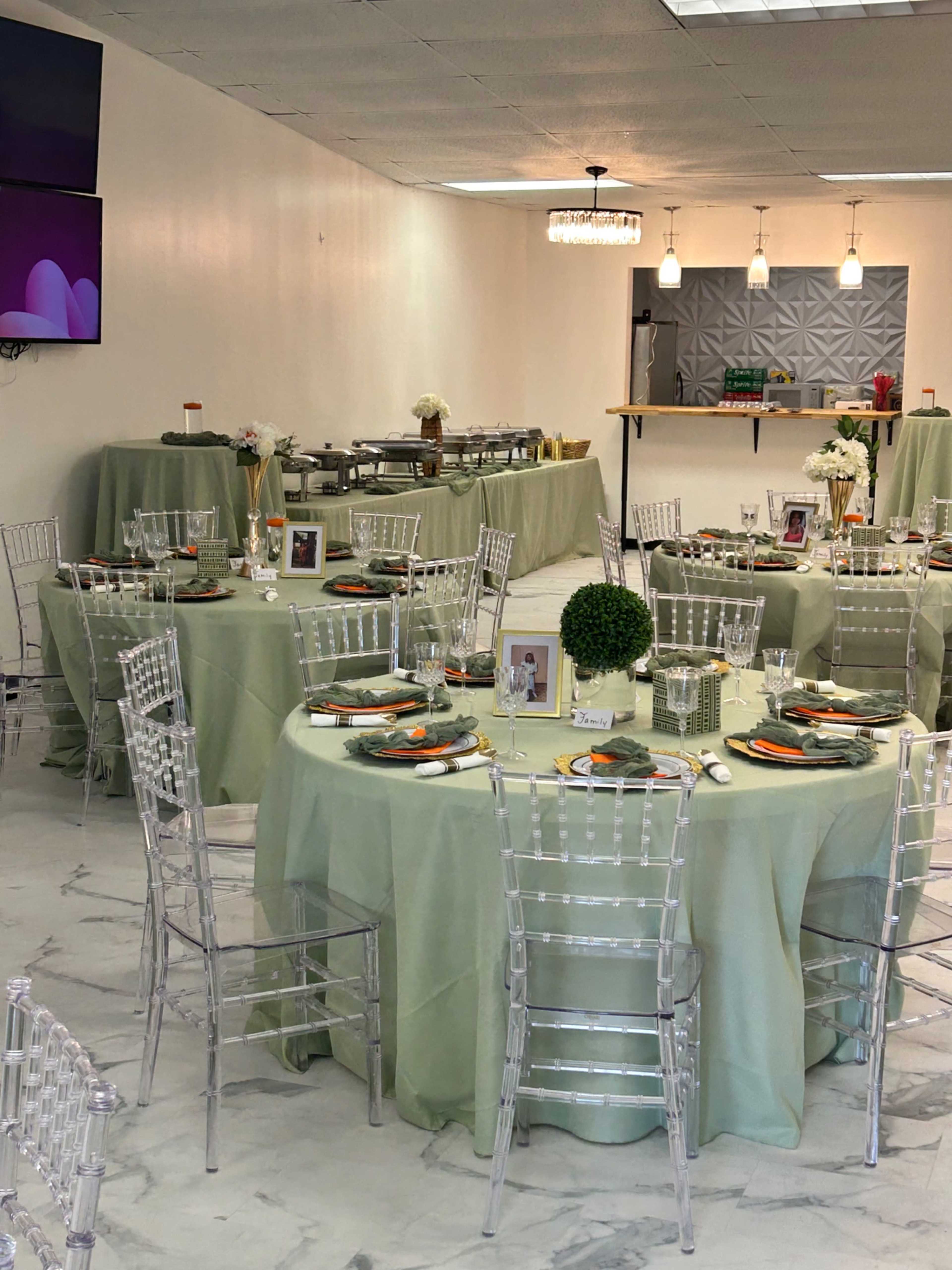A spacious dining area features round tables dressed in green tablecloths, set with elegant dishware and centerpieces, while a buffet station and a wall-mounted television are in the background.