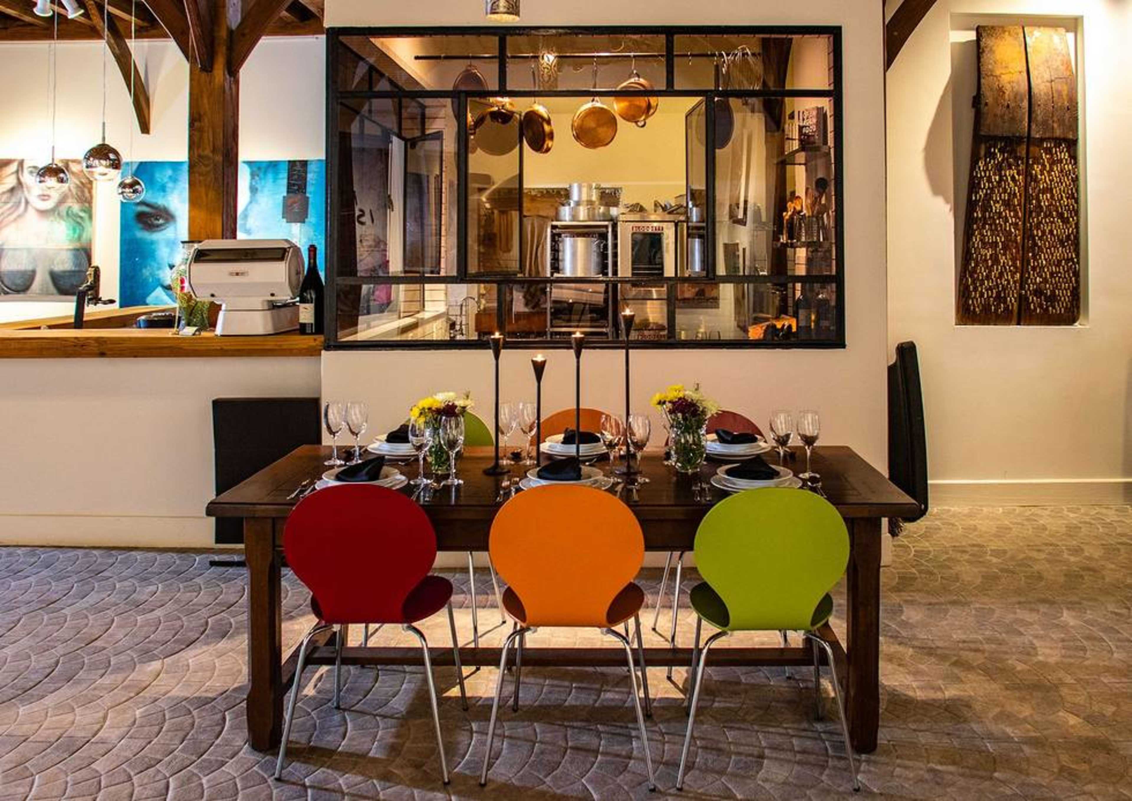 A wooden dining table is set with plates and glassware, surrounded by colorful chairs in an interior that features a view into a kitchen area with hanging pots.