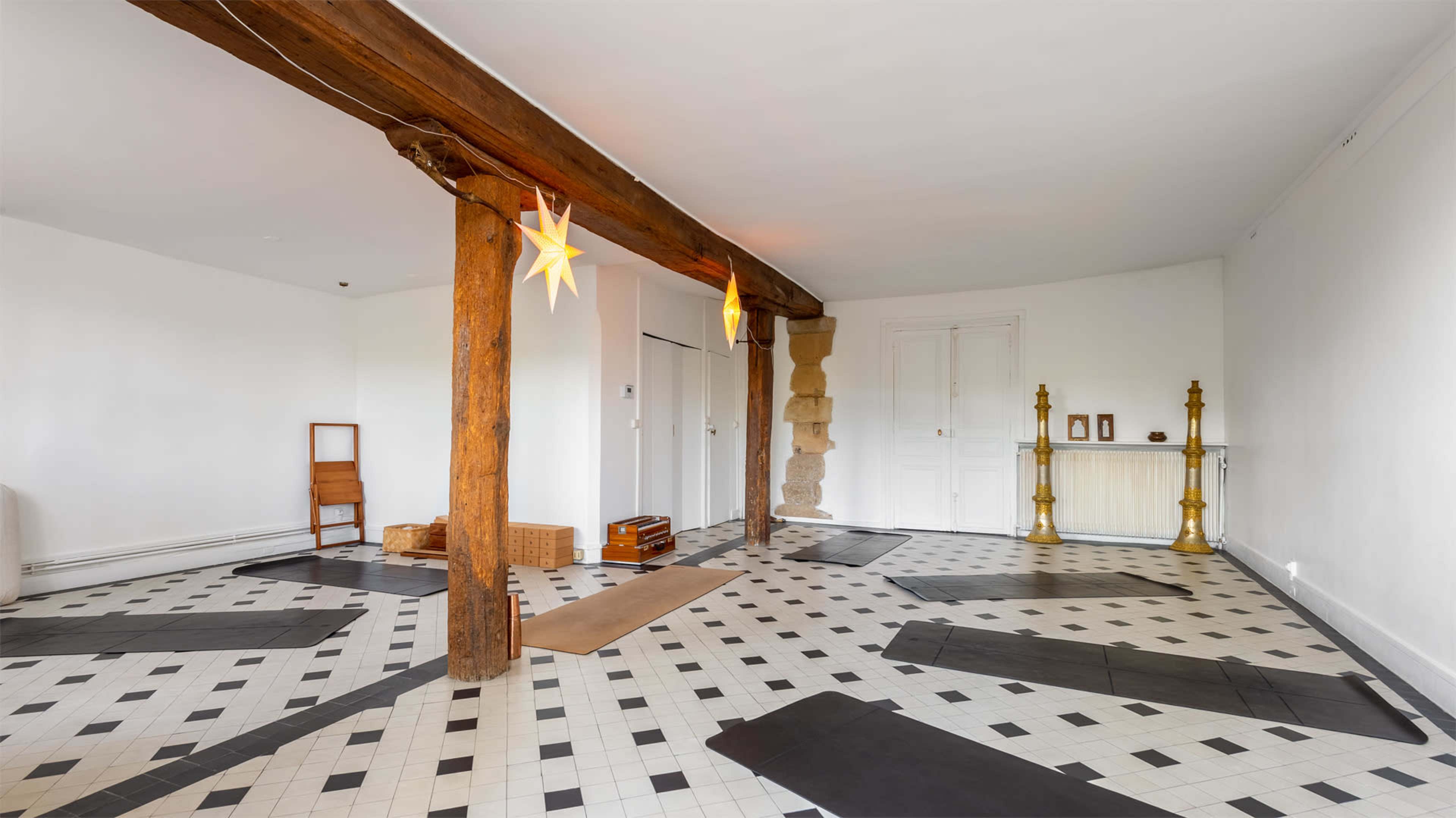 A spacious, minimally furnished room with wooden beams, black and white tile flooring, and several yoga mats arranged for use.