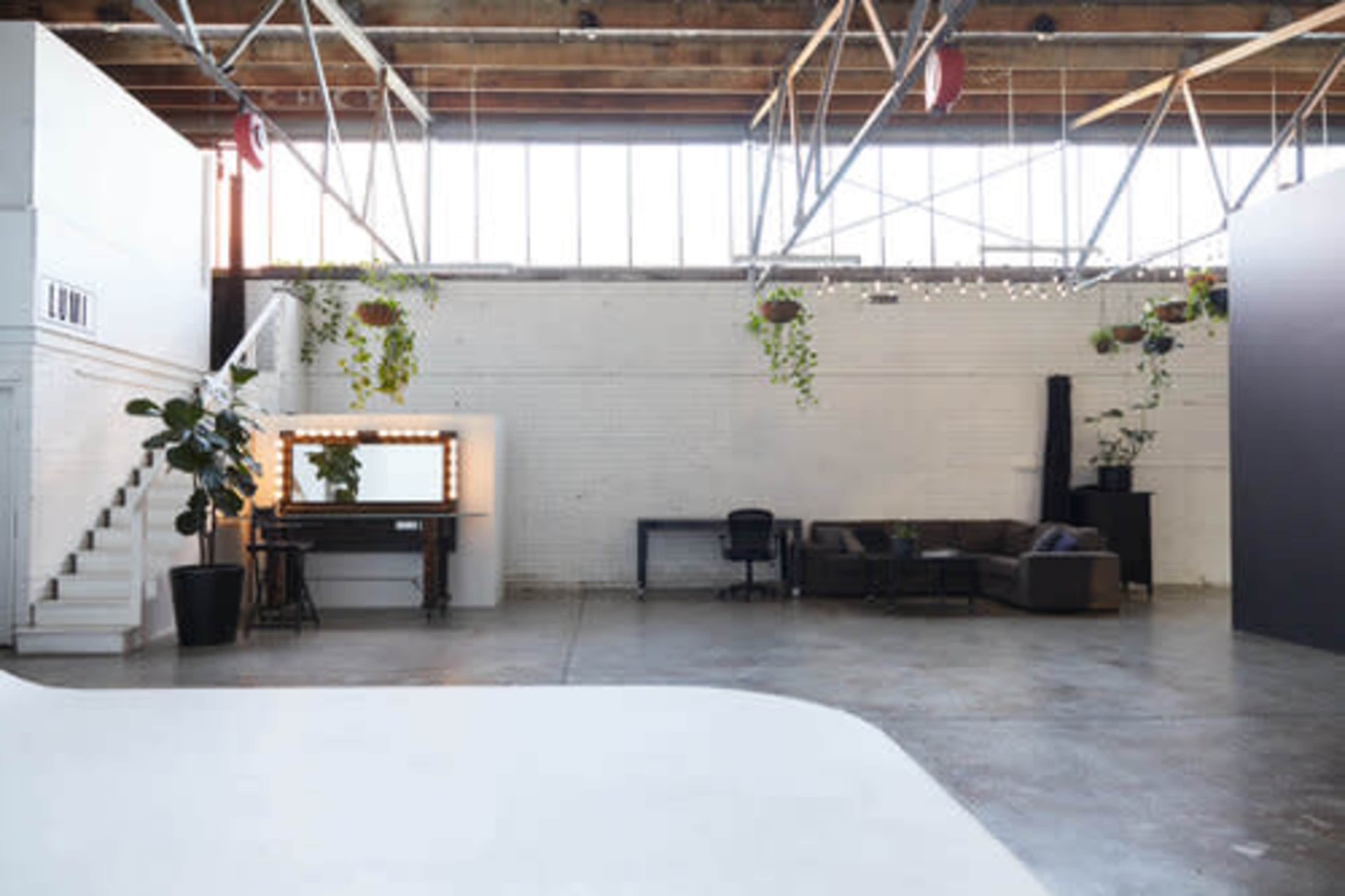A spacious industrial-style room with white brick walls, large windows, a staircase, a makeup vanity with lights, a sofa, and a desk.