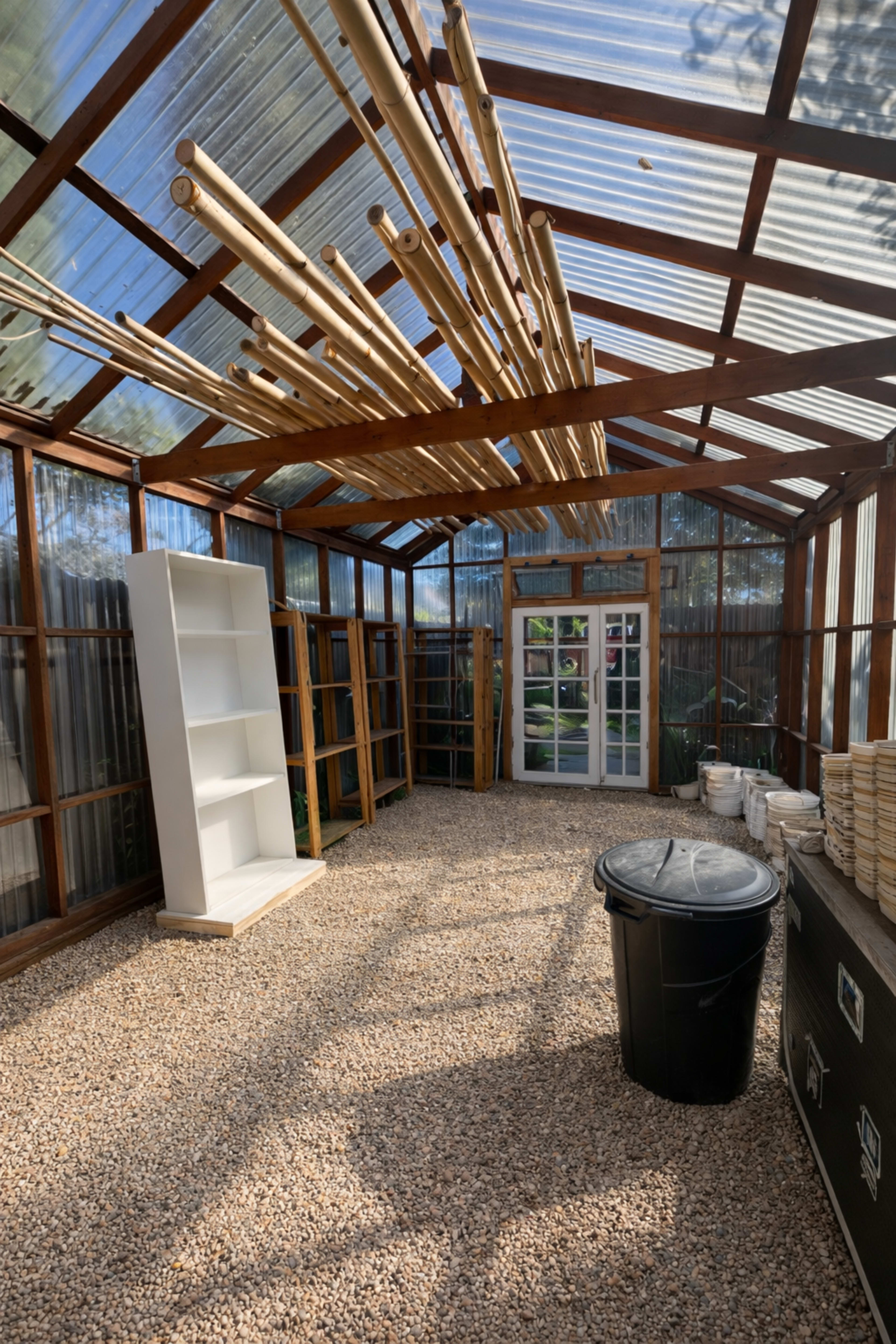 The interior of a greenhouse features gravel flooring, wooden shelving units, and a series of bamboo rods suspended from the ceiling.