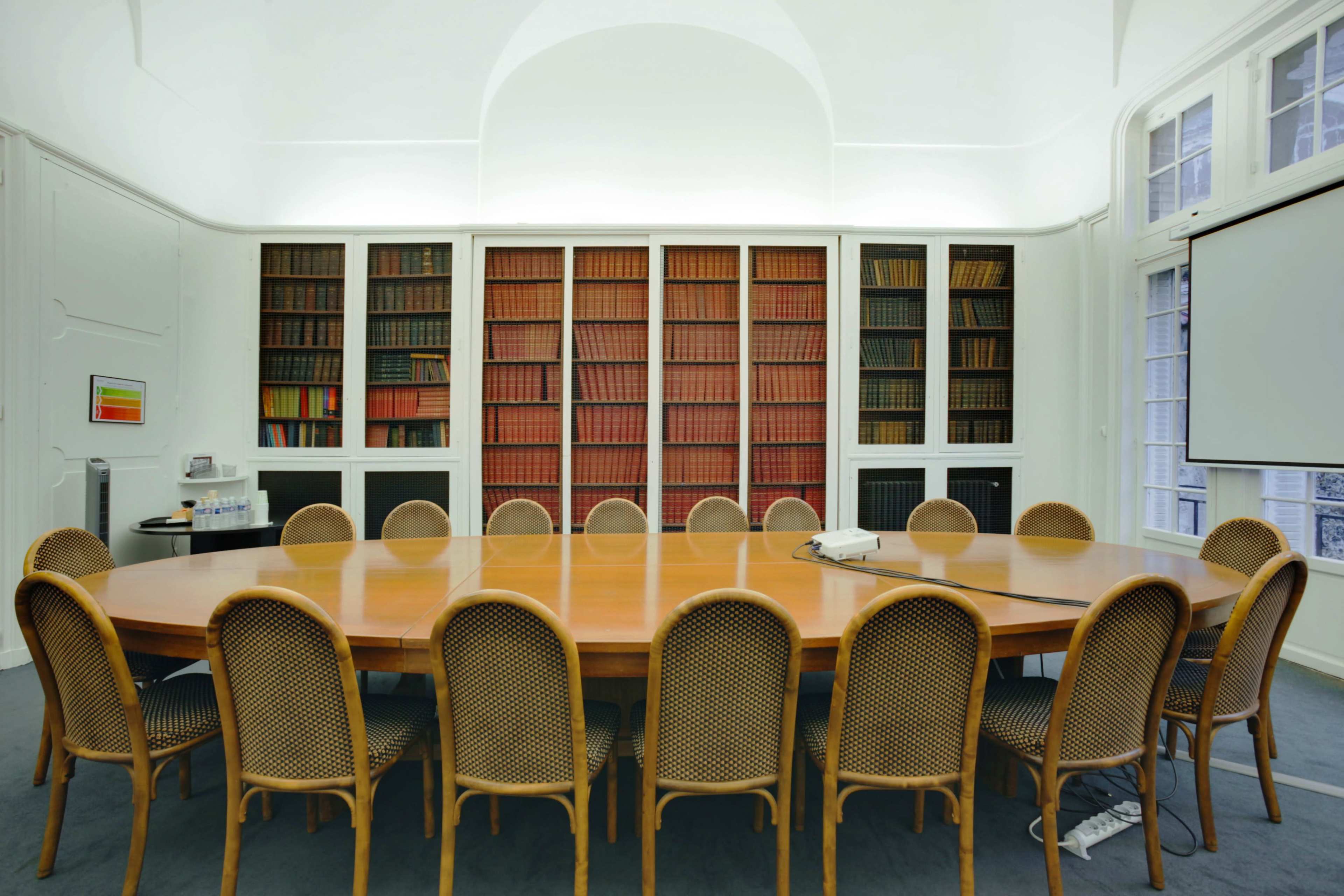 A spacious conference room features a large round wooden table surrounded by thirteen cane-back chairs, with shelves of books lining the walls.