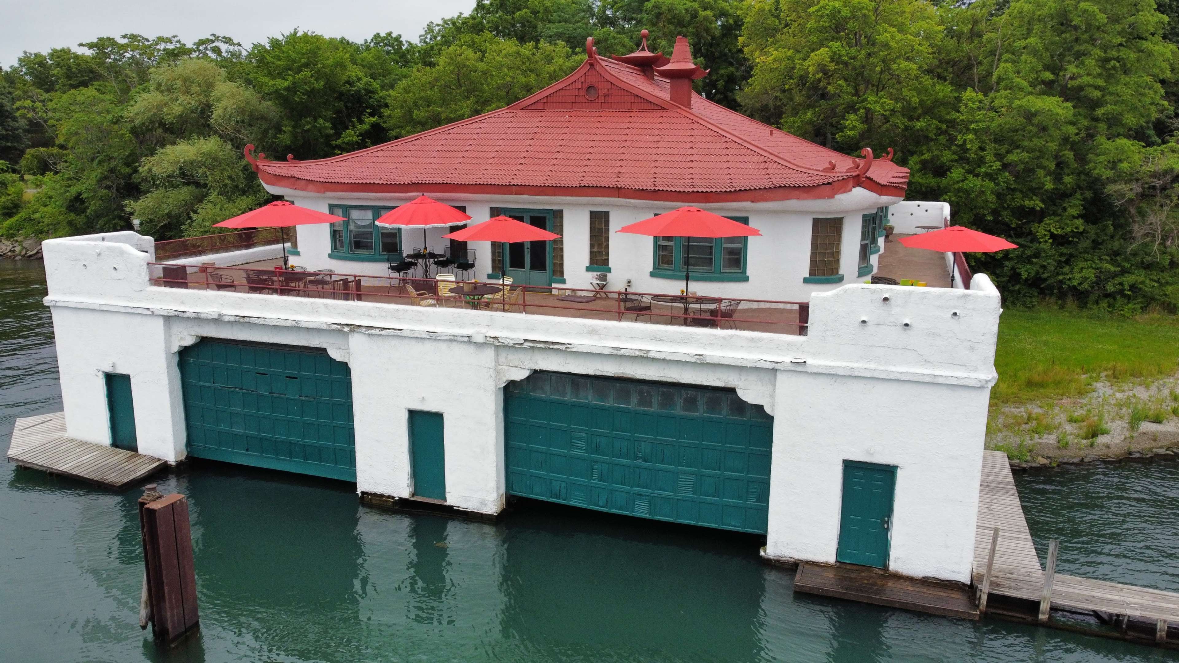 The image shows a white building with a red-tiled roof and large green doors, situated on the water and featuring red umbrellas over outdoor seating.