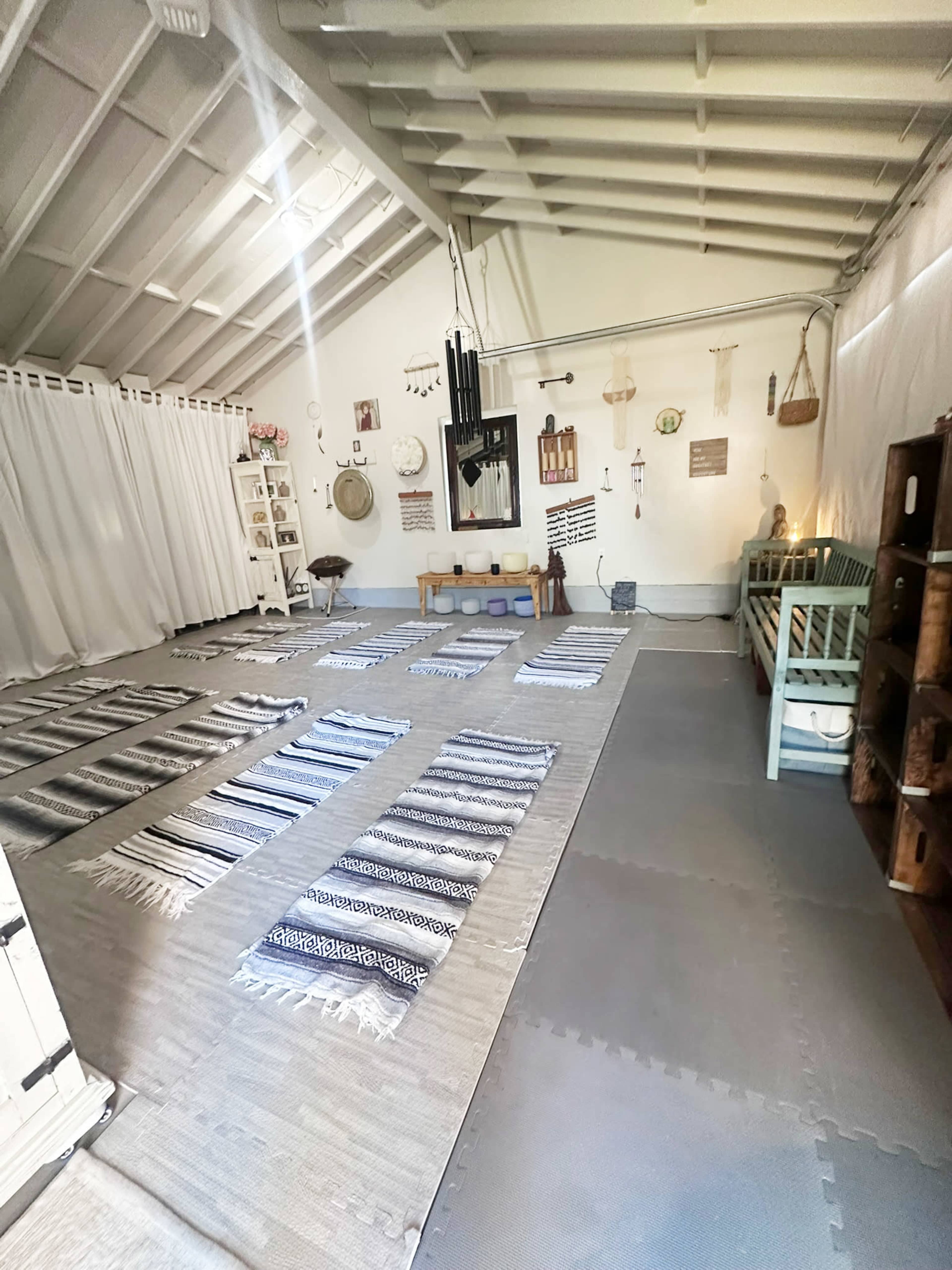 The room features a spacious layout with several patterned rugs arranged on a neutral floor, surrounded by simple wooden furniture and decor items.