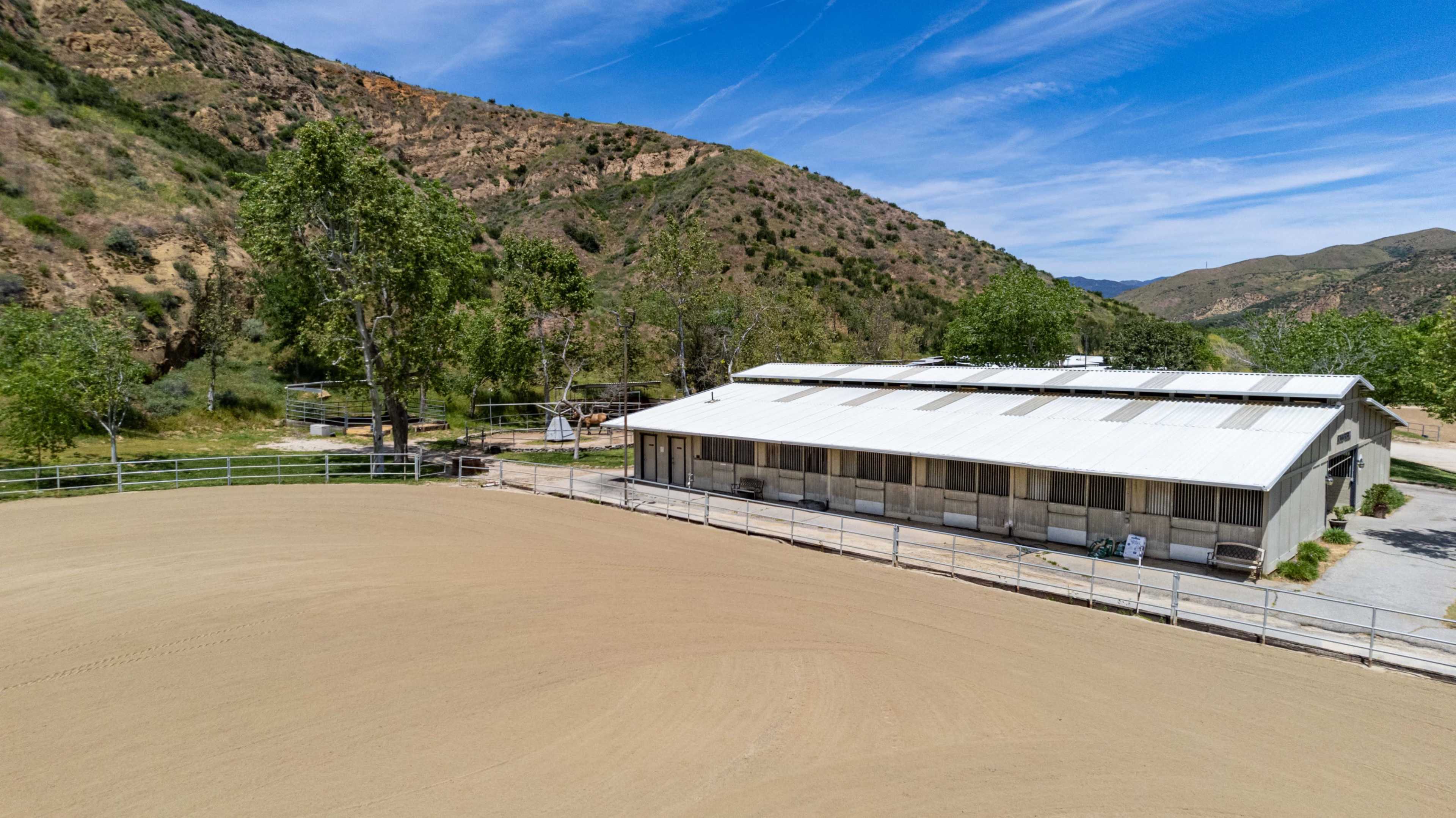 A metal building stands beside a sandy horse arena, enclosed by a fence and surrounded by hills and trees.