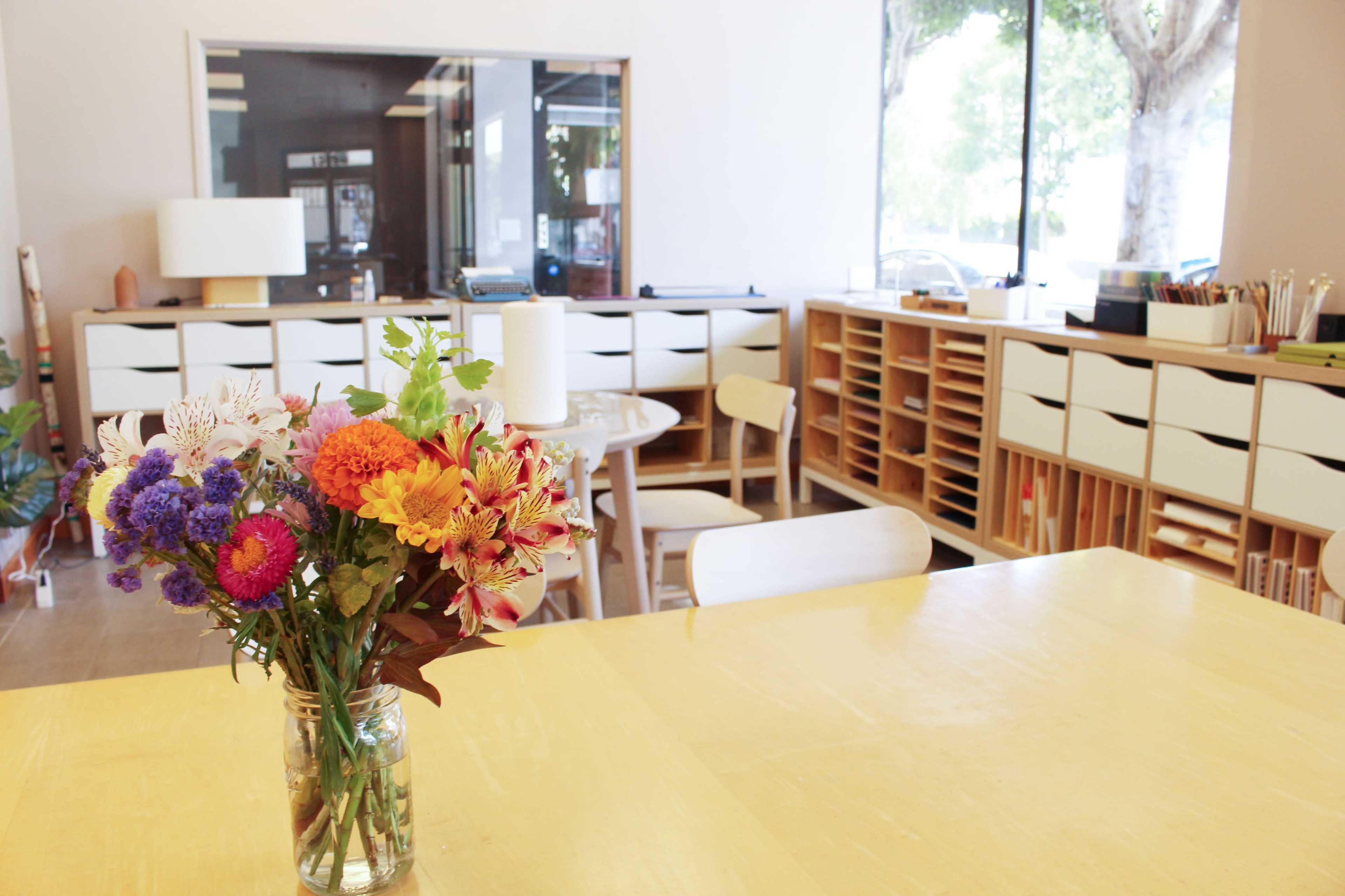 A bouquet of colorful flowers sits in a glass jar on a yellow table in a bright, organized workspace with wooden shelves and chairs.