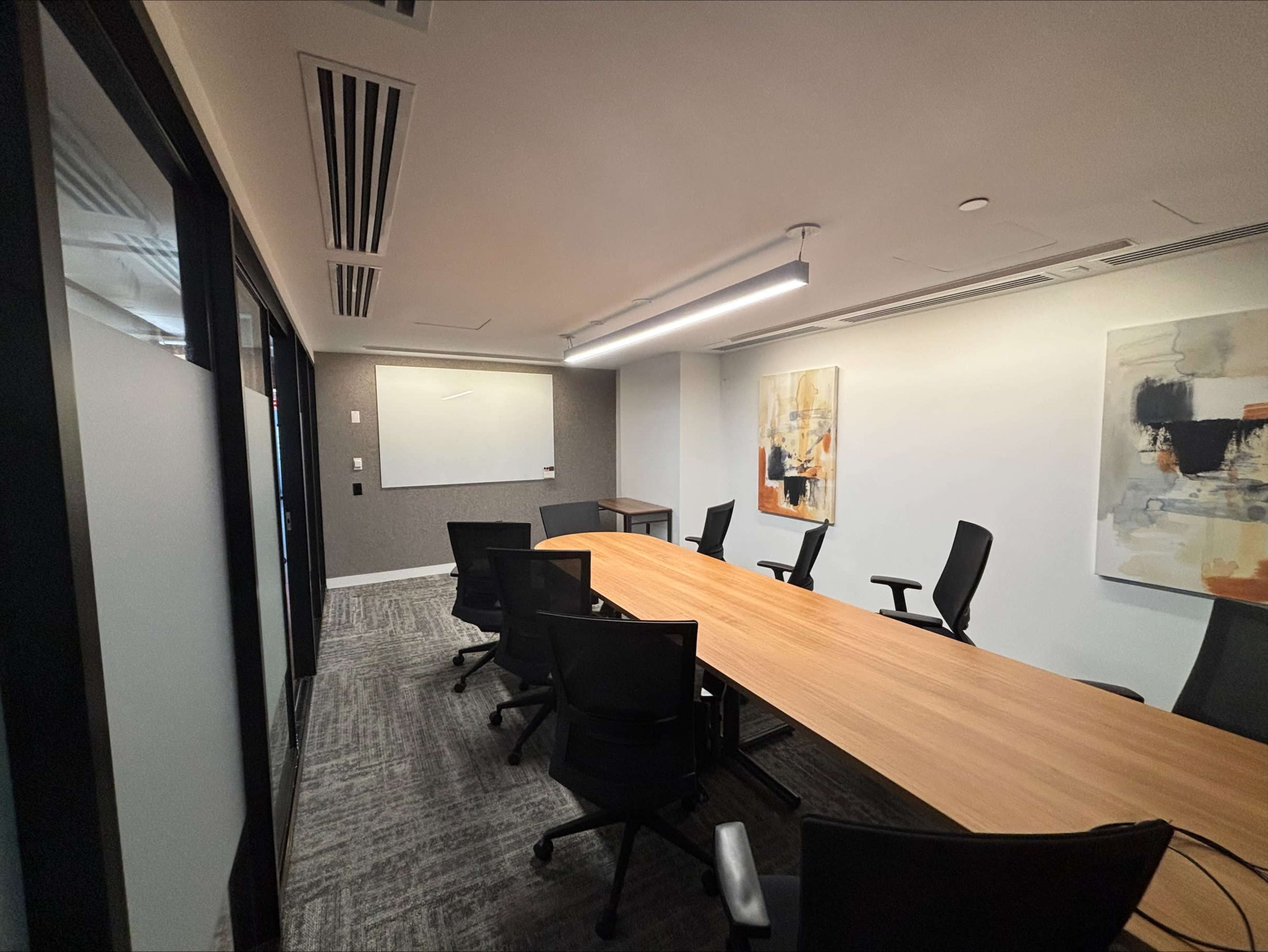 The image shows a modern conference room featuring a long wooden table surrounded by black office chairs, with a whiteboard and two abstract paintings on the walls.