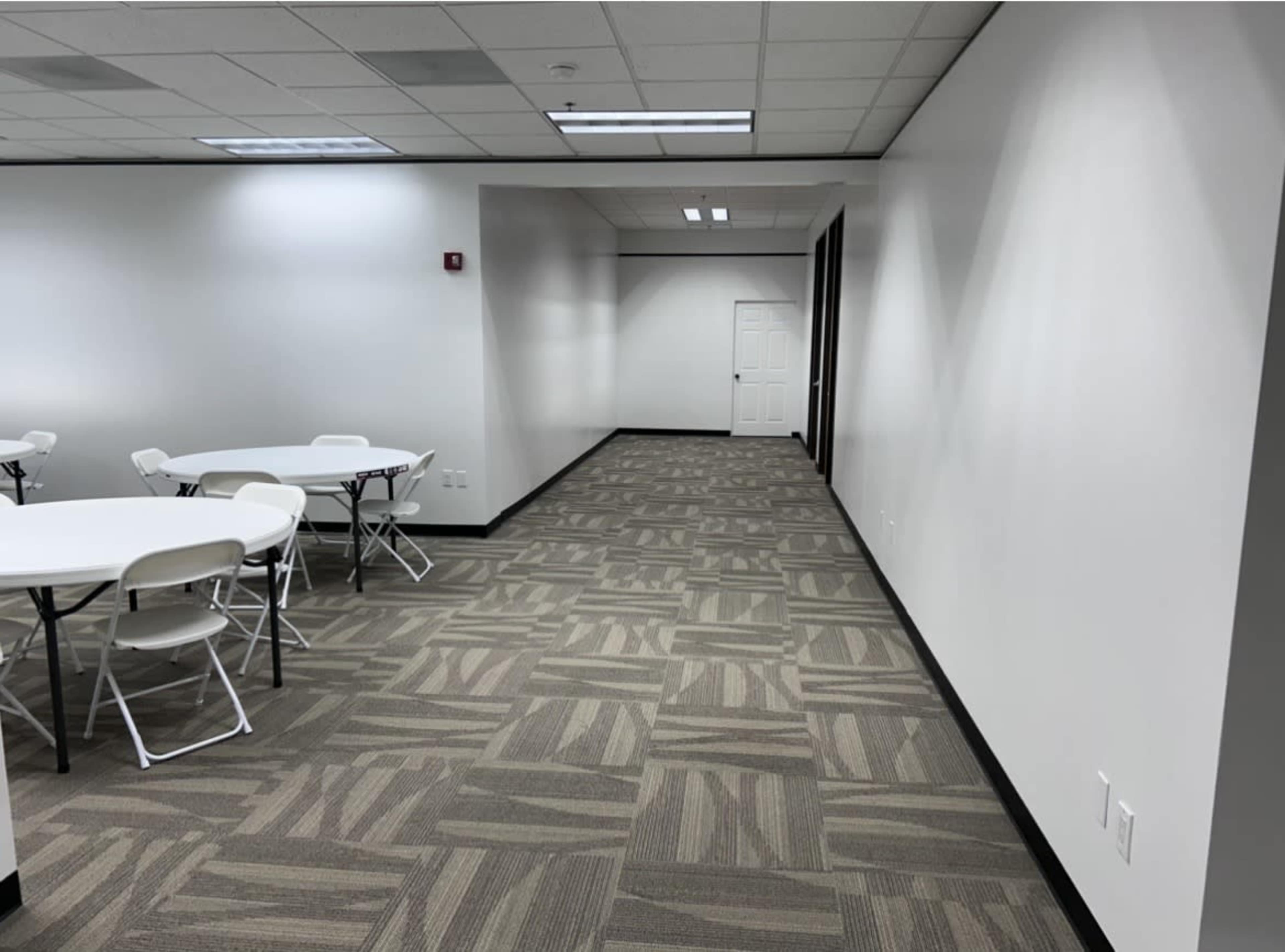 The image shows a bright, empty room featuring white walls, a tan carpet with a geometric pattern, and several round tables with white folding chairs arranged around them, leading to a hallway with a closed door at the end.