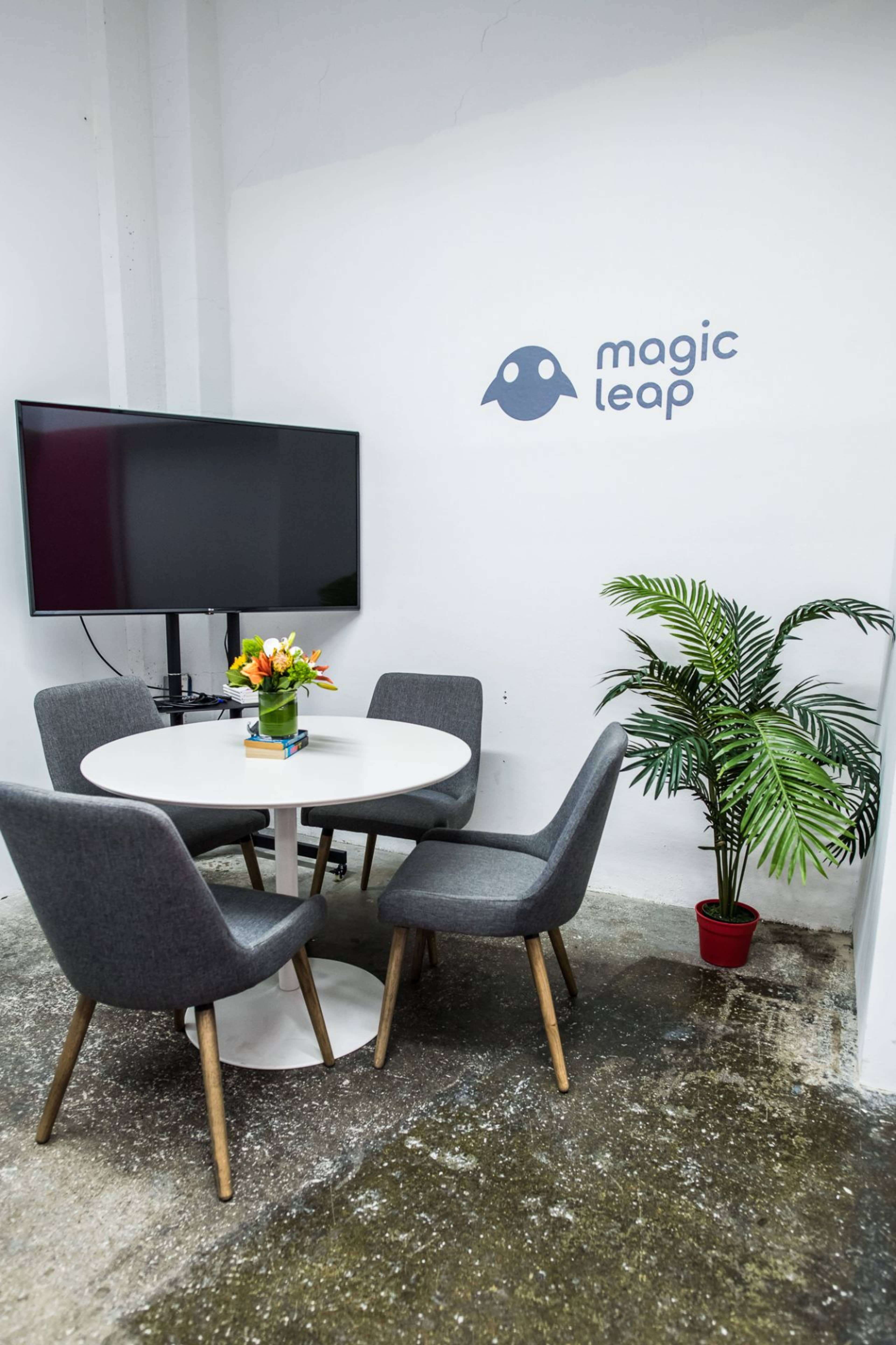 A modern meeting room featuring a round table with four grey chairs, a television on the wall, a potted plant, and a wall displaying the "magic leap" logo.