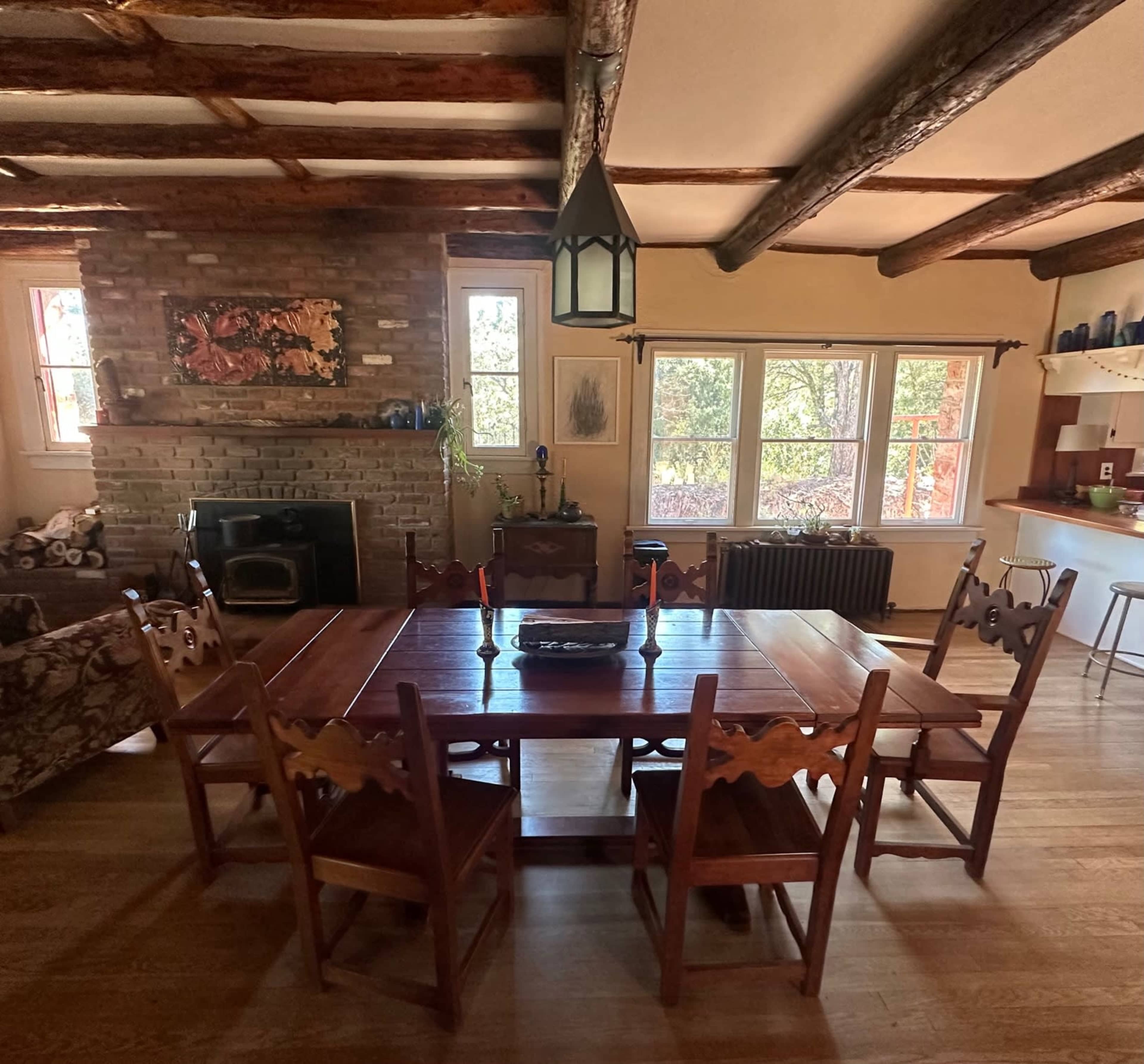 A rustic dining area with a large wooden table surrounded by six chairs, a brick fireplace, and windows overlooking a wooded view.