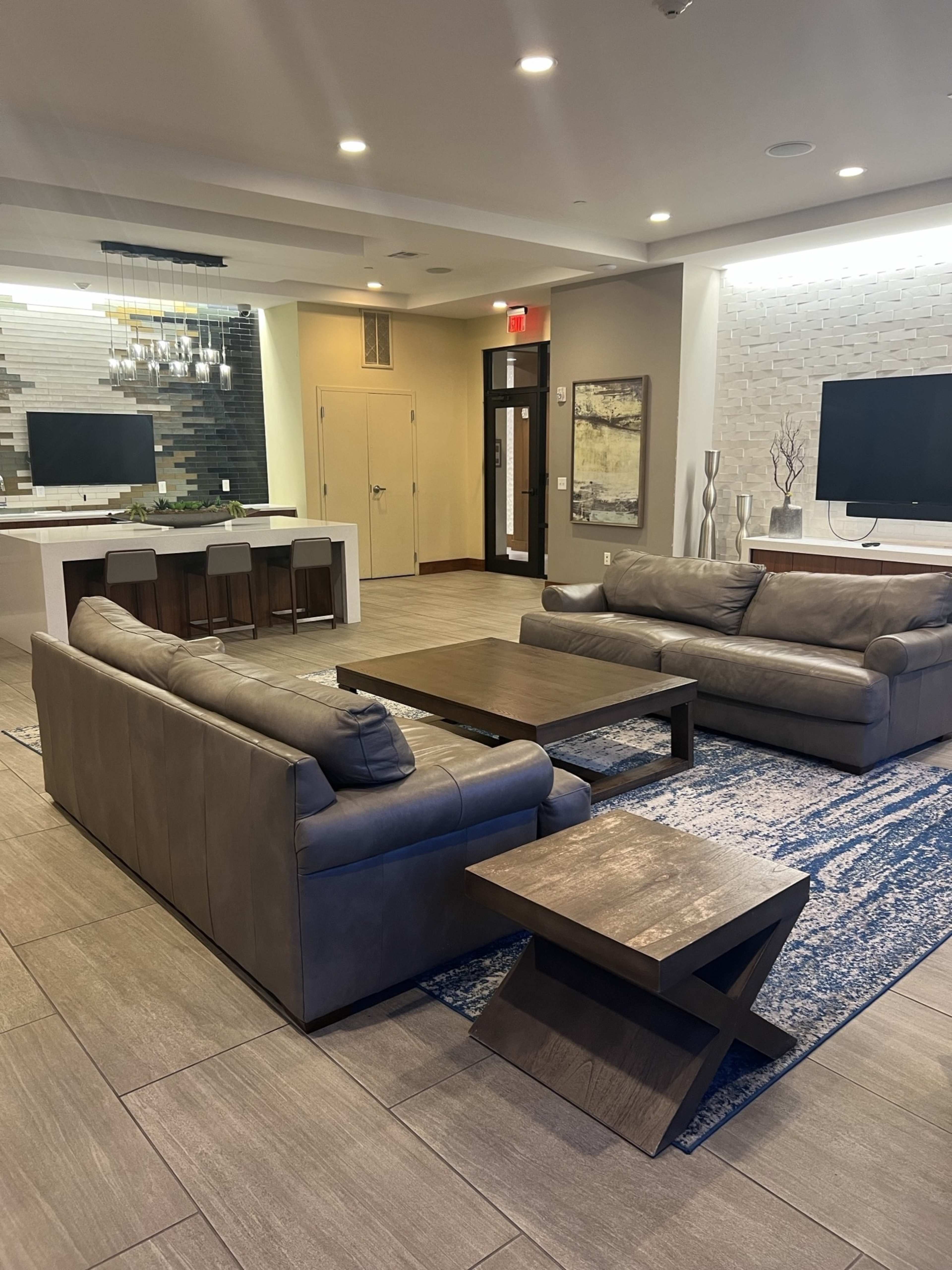 The image shows a modern lounge area featuring two gray leather sofas, a wooden coffee table, and a small side table, all set on a blue and white patterned rug.