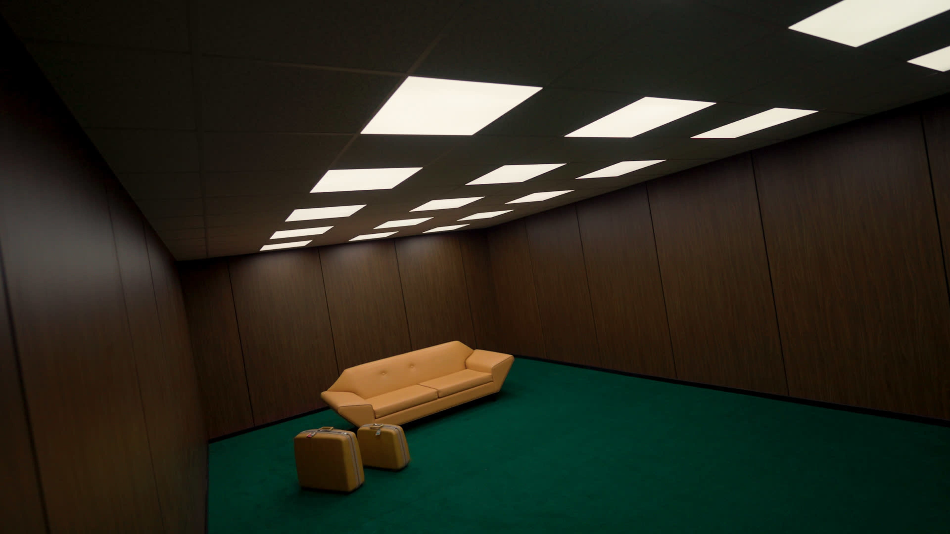 The image shows a sparsely furnished room with wood-paneled walls, a green carpet, a yellow couch, and two light-colored suitcases.