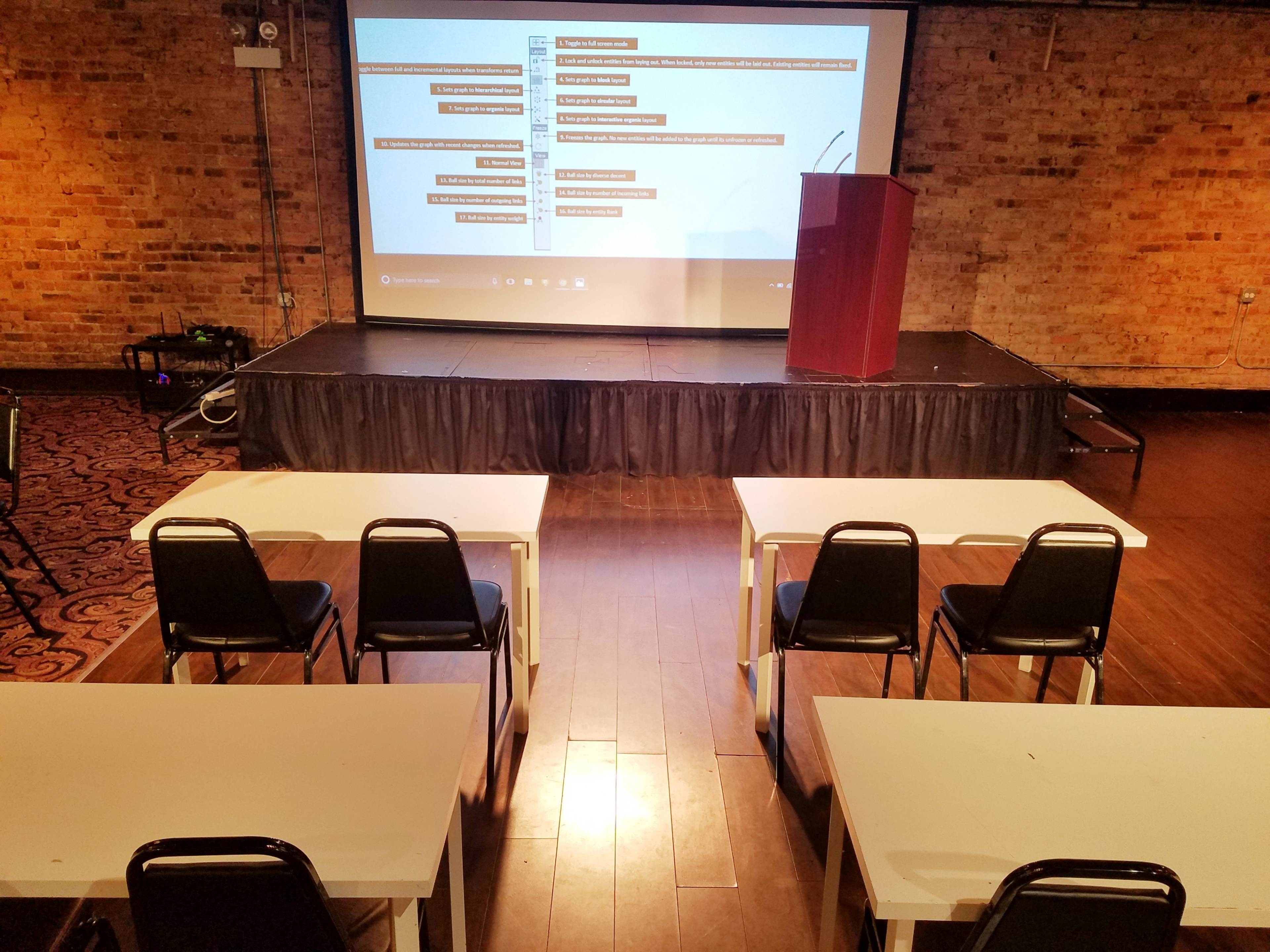 The image shows a presentation setup in a room with a stage, a podium, and several rows of empty tables and chairs facing a screen displaying content.