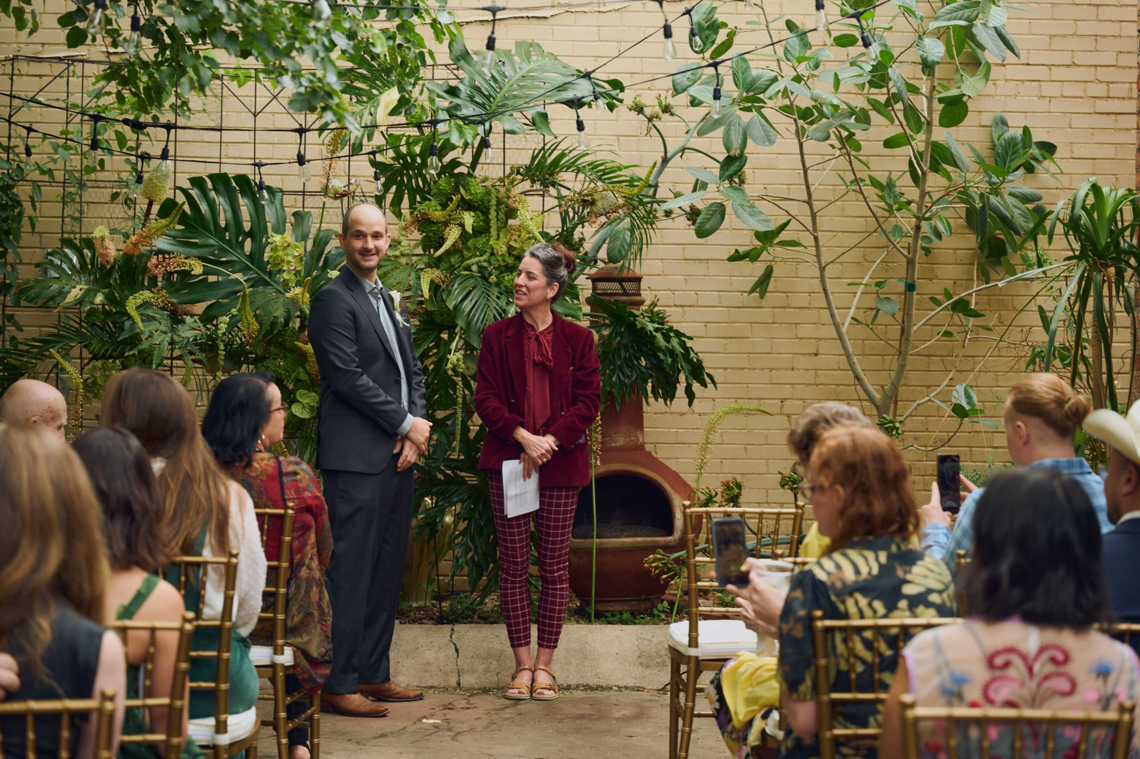 A couple stands in front of an audience in a lush, green outdoor setting, preparing for a ceremony.