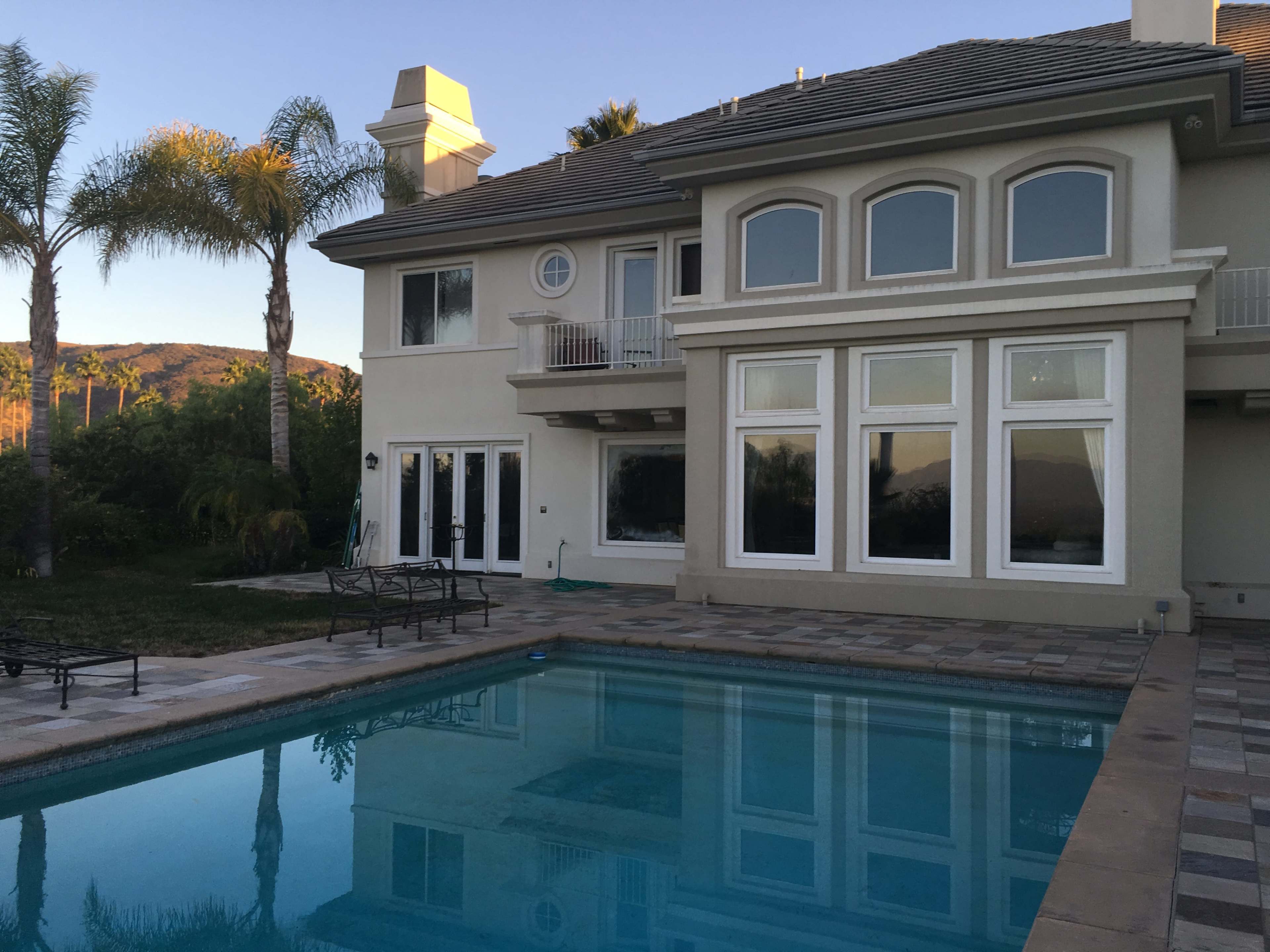A two-story house with large windows, a swimming pool in the foreground, and palm trees in the background.