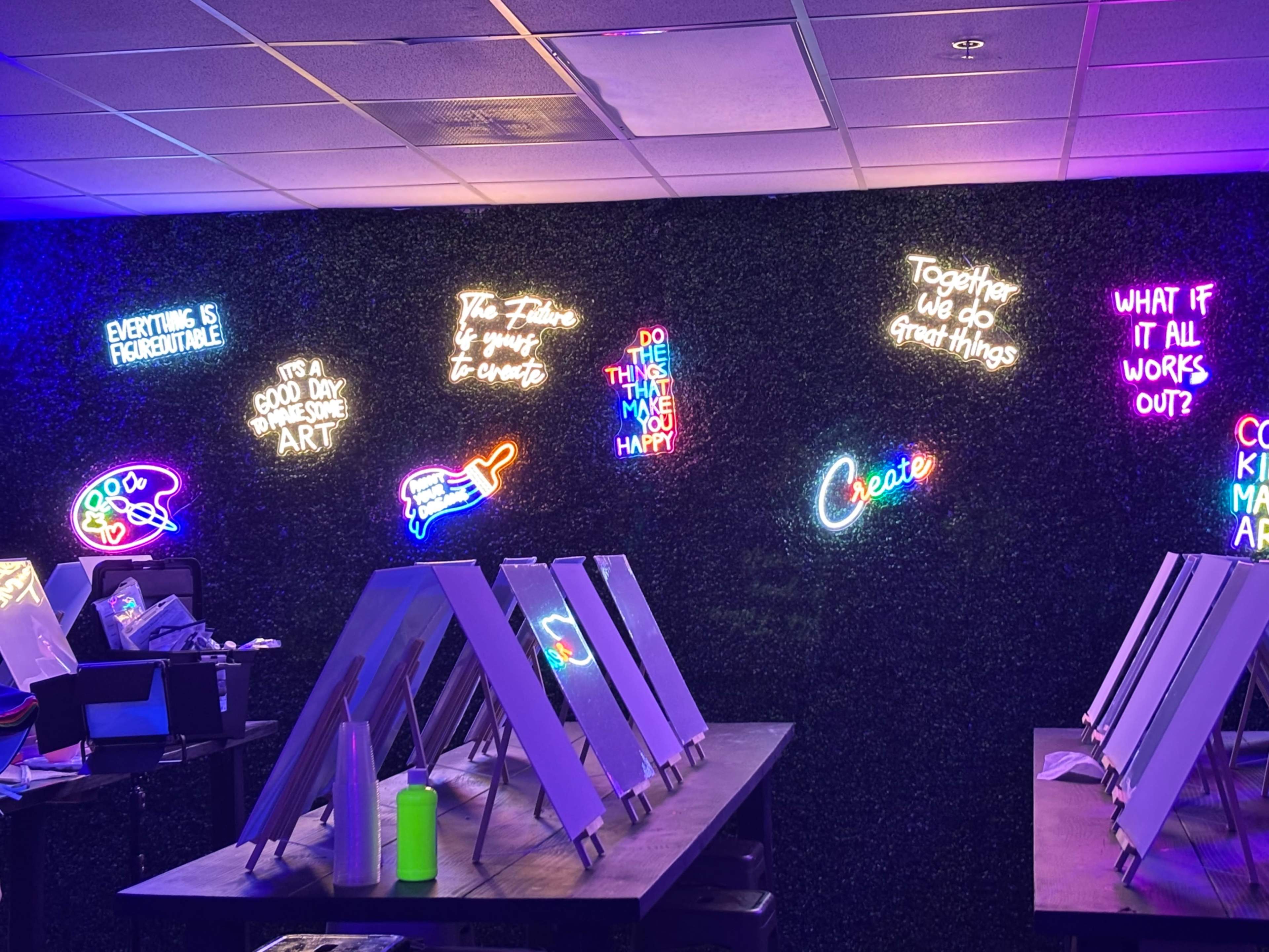 The image shows a vibrant indoor setting with neon signs on a green wall and several easels on tables.