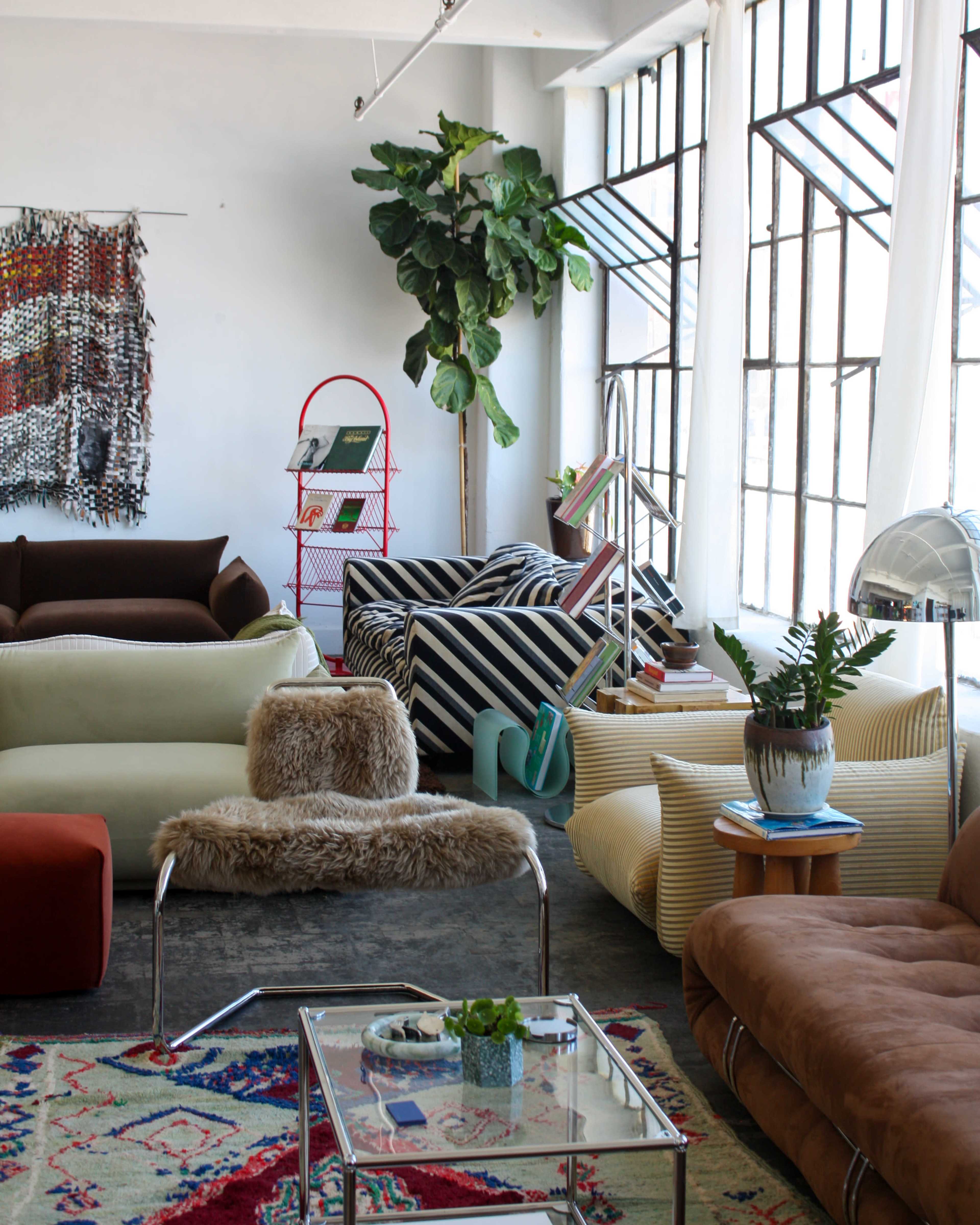 The image shows a large, well-lit living room with various styles of couches, a potted plant, a glass coffee table, and shelves with books.
