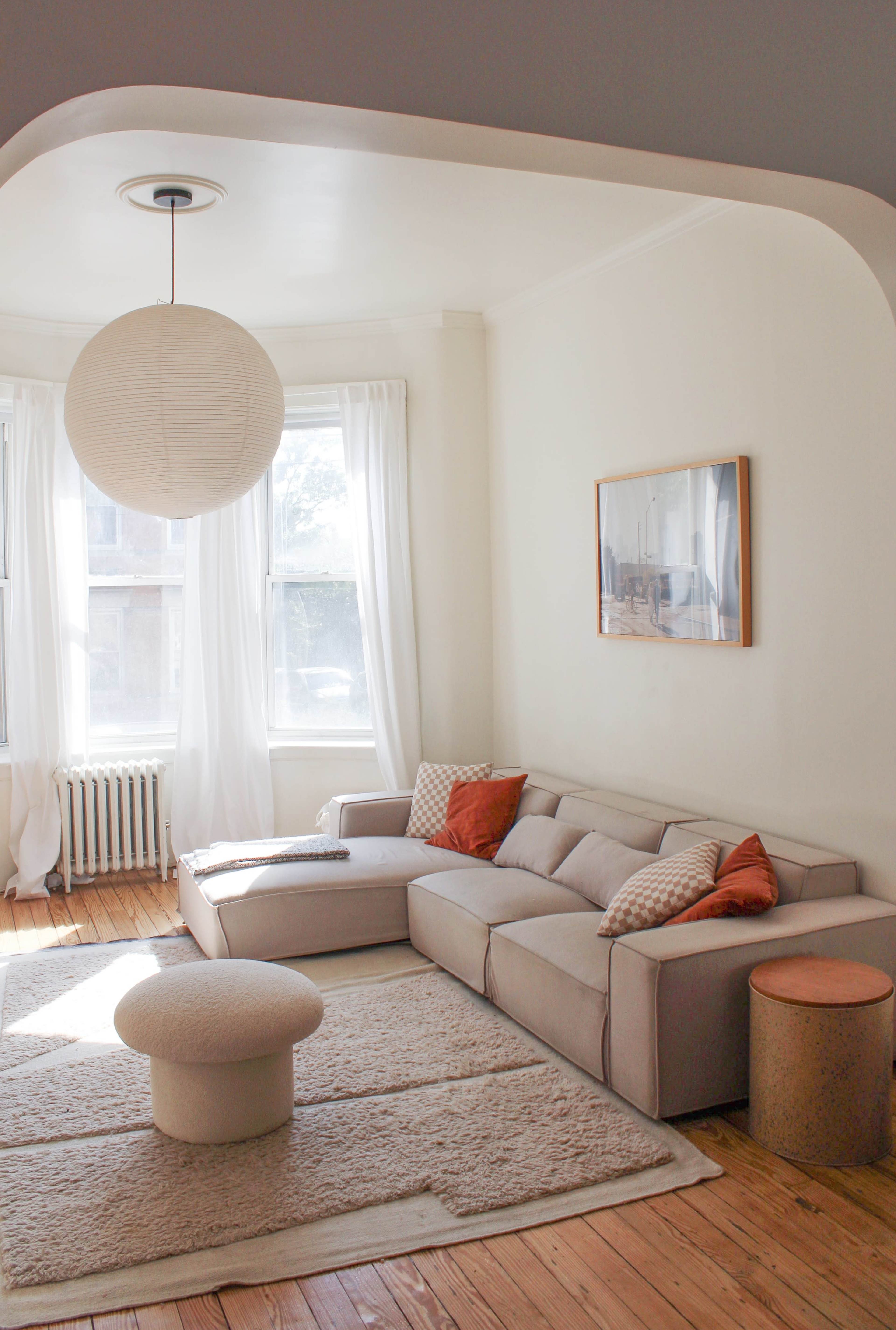The image shows a bright living room with a light-colored sectional sofa, a circular coffee table, and large windows with sheer curtains.