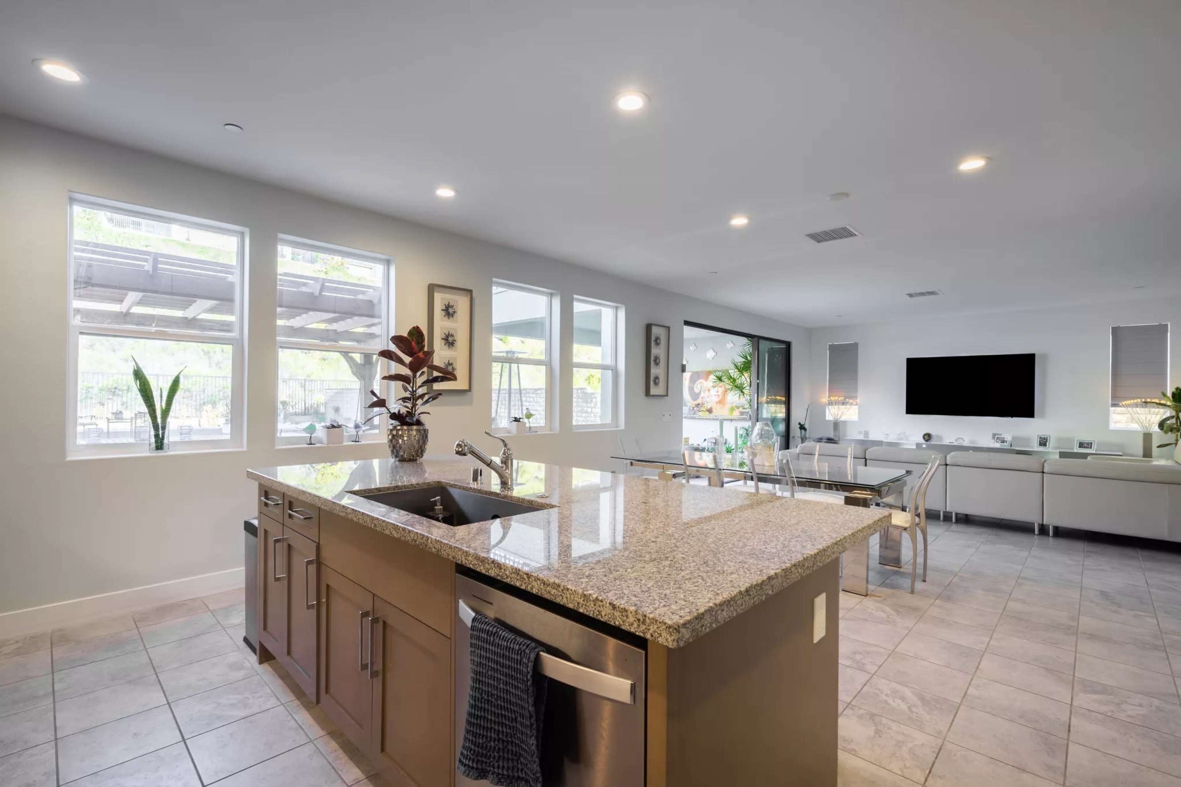 A modern kitchen with a granite countertop, featuring a sink and appliances, adjacent to a dining area with large windows and natural light.