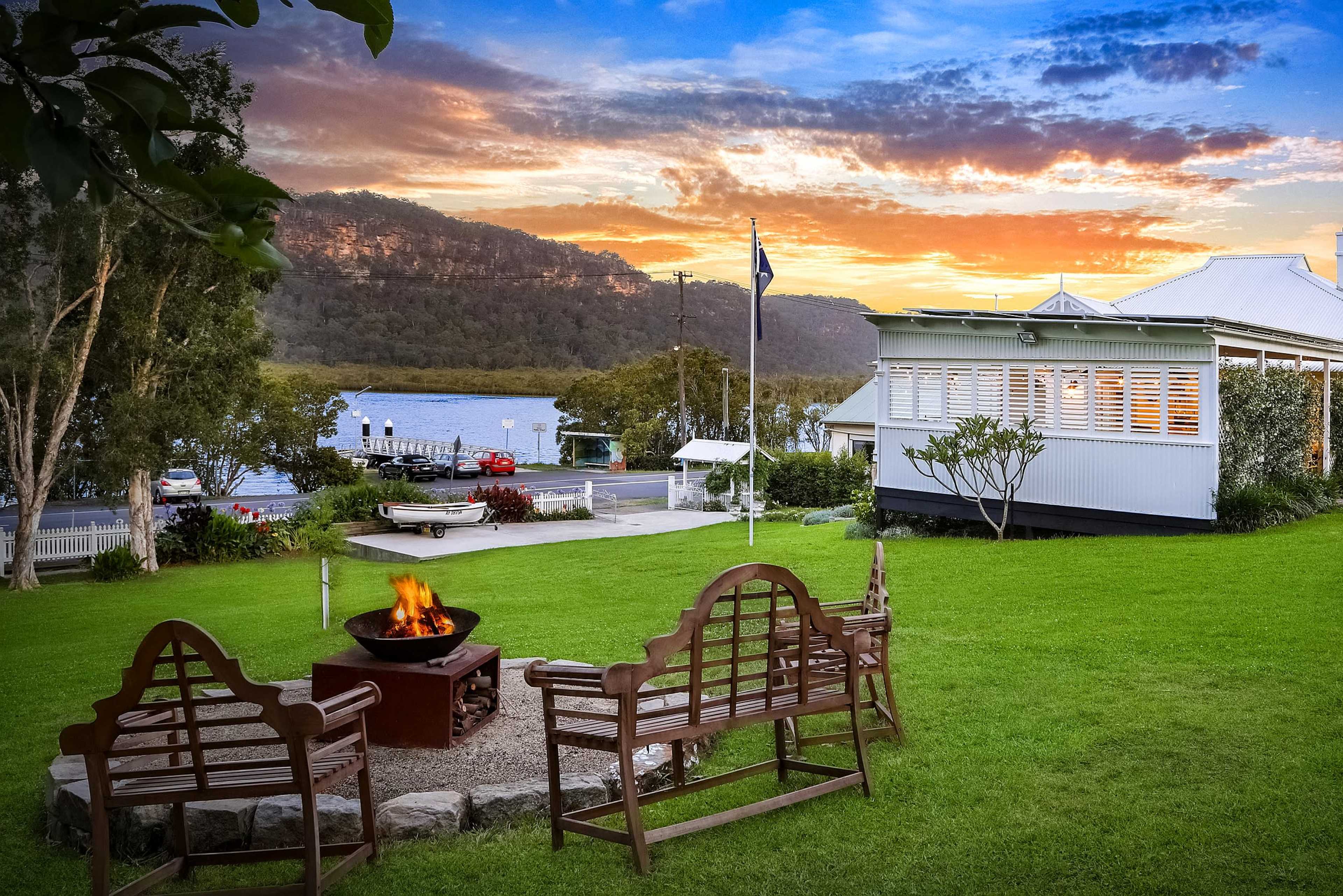 A fire pit is surrounded by wooden benches on a lawn with a view of a river and a sunset in the background.