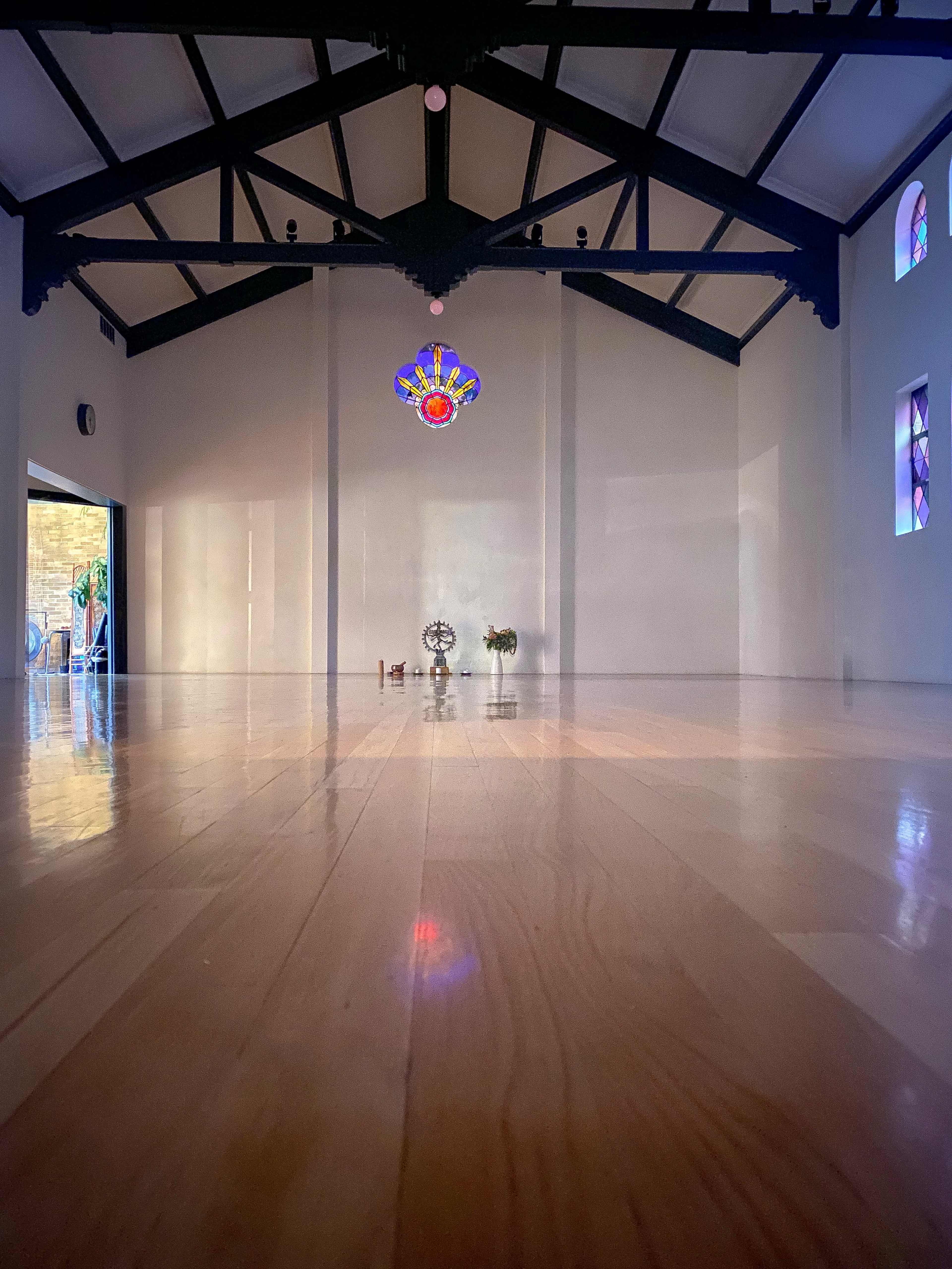 The image depicts a spacious, empty room with high ceilings, wooden flooring, and a colorful stained glass feature at the top.