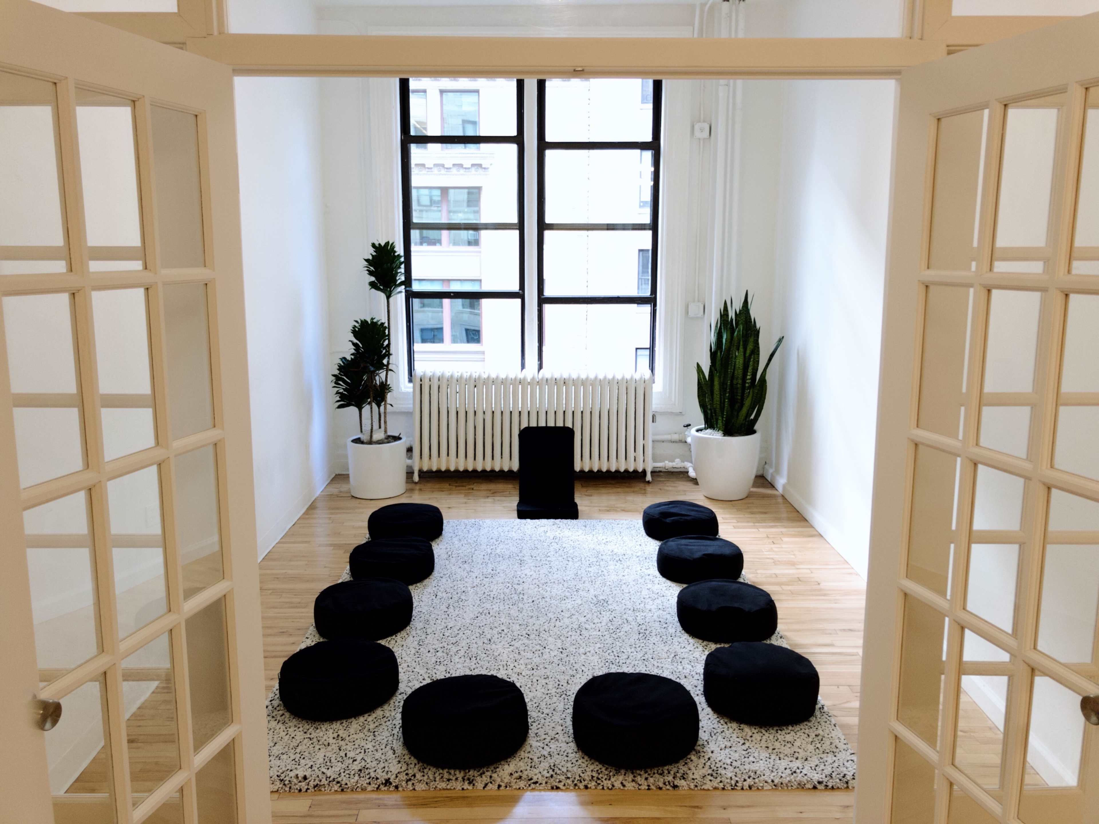 A spacious, bright room with a circular arrangement of black cushions on a light-colored rug, flanked by plants and a radiator below large windows.