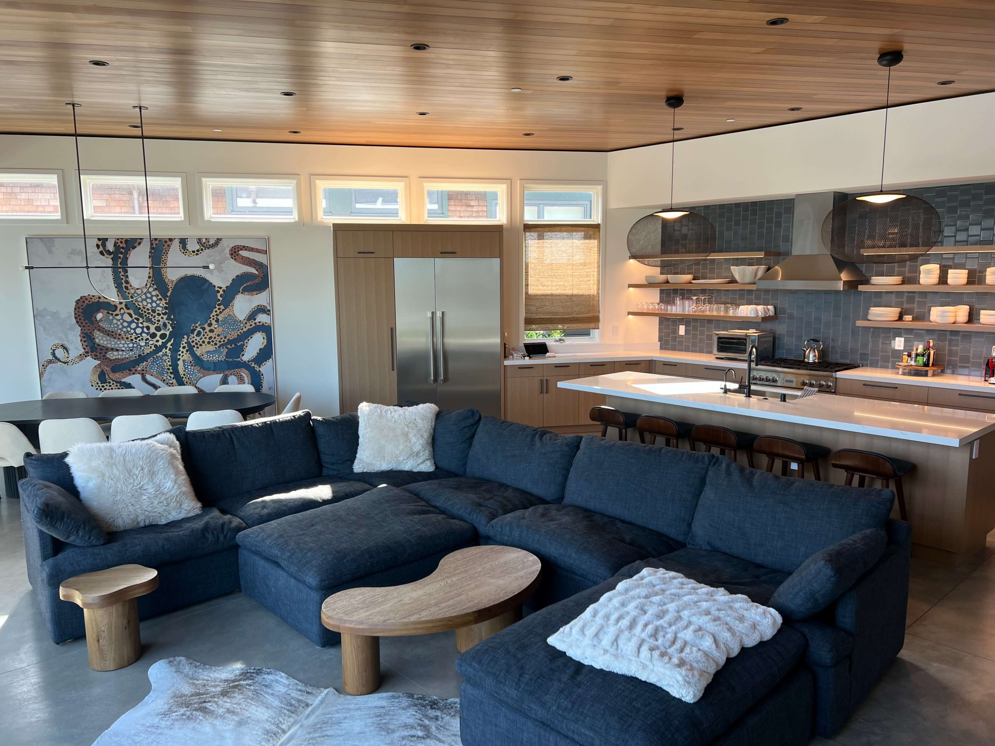 The image shows a modern living space featuring a large sectional sofa, a wooden coffee table, and an open kitchen with stylish cabinetry and a wall art piece depicting an octopus.