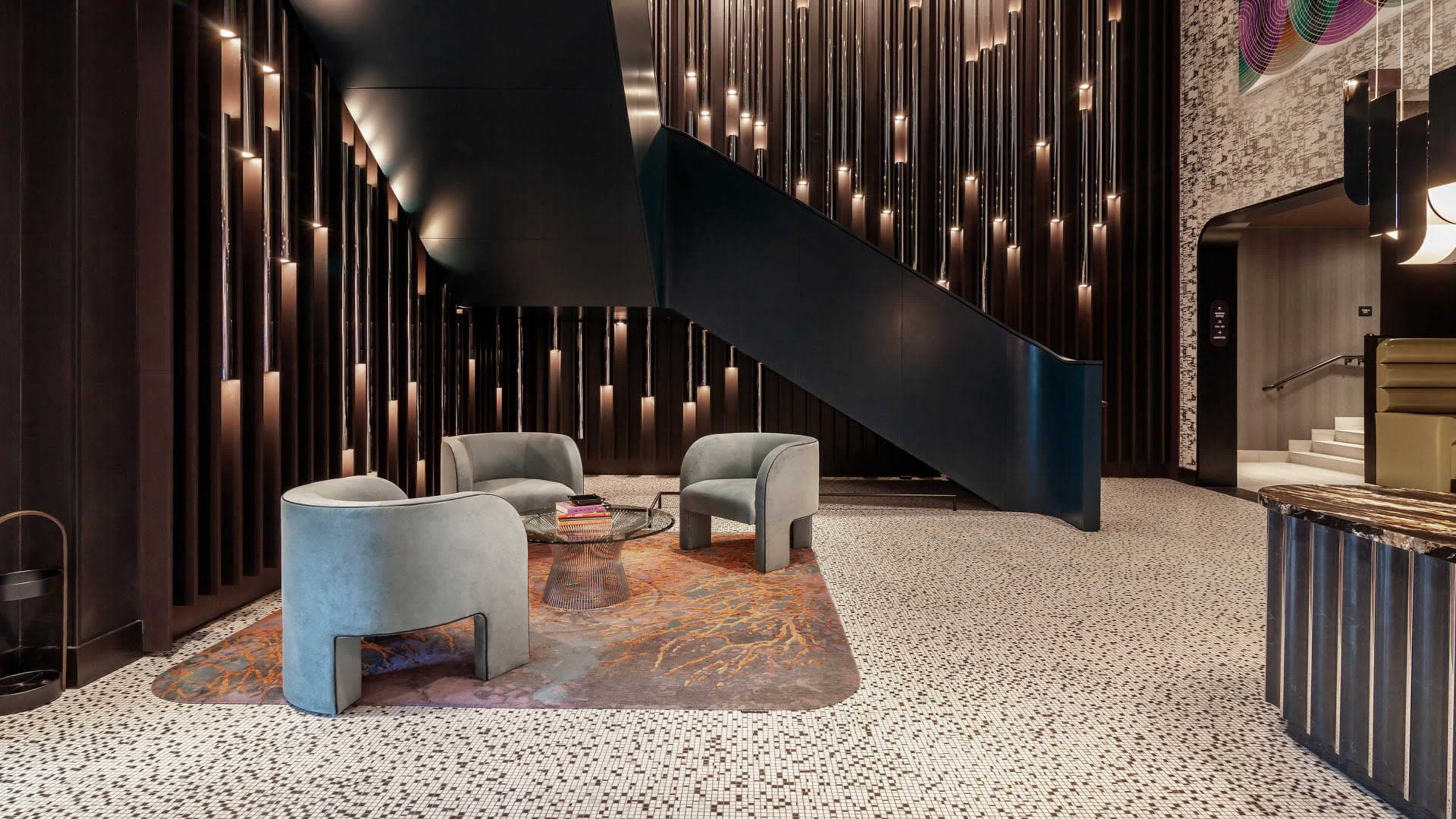 The image shows a modern lobby with a staircase, featuring textured walls, a patterned floor, and circular seating arrangements.