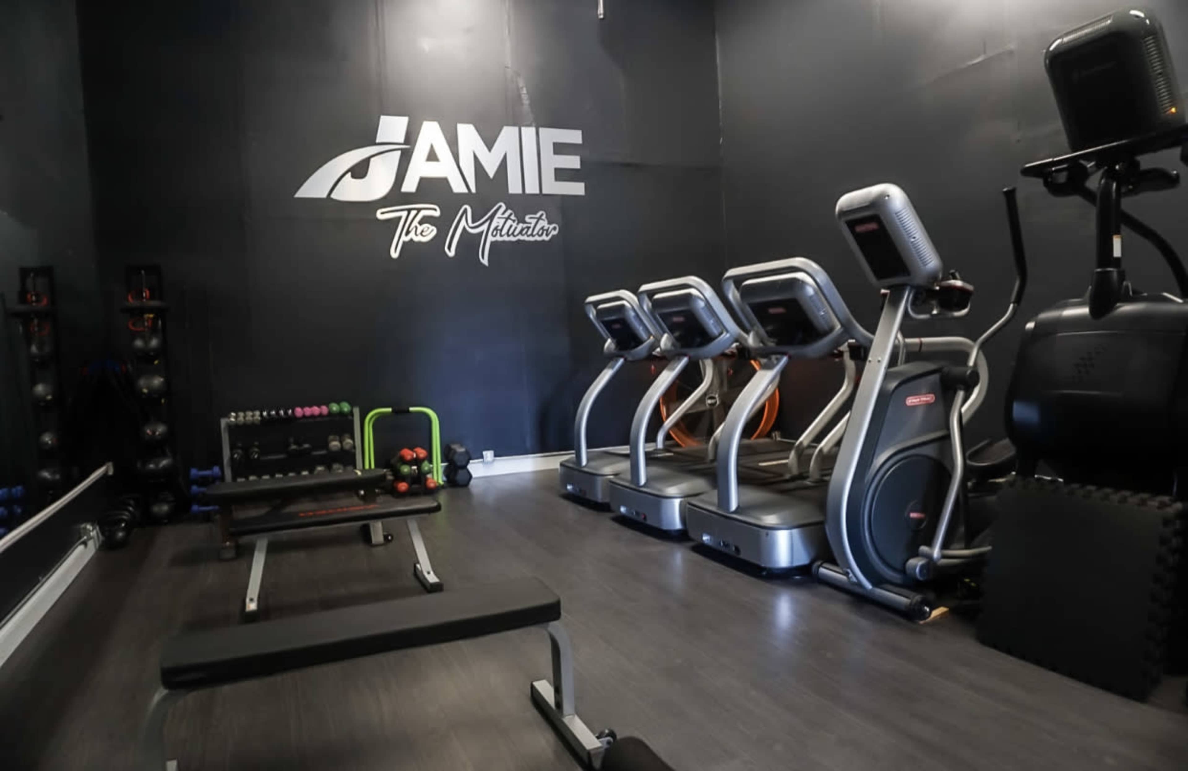 A small gym with multiple elliptical machines, a weight bench, and various dumbbells against a dark wall with the name "Jamie" prominently displayed.