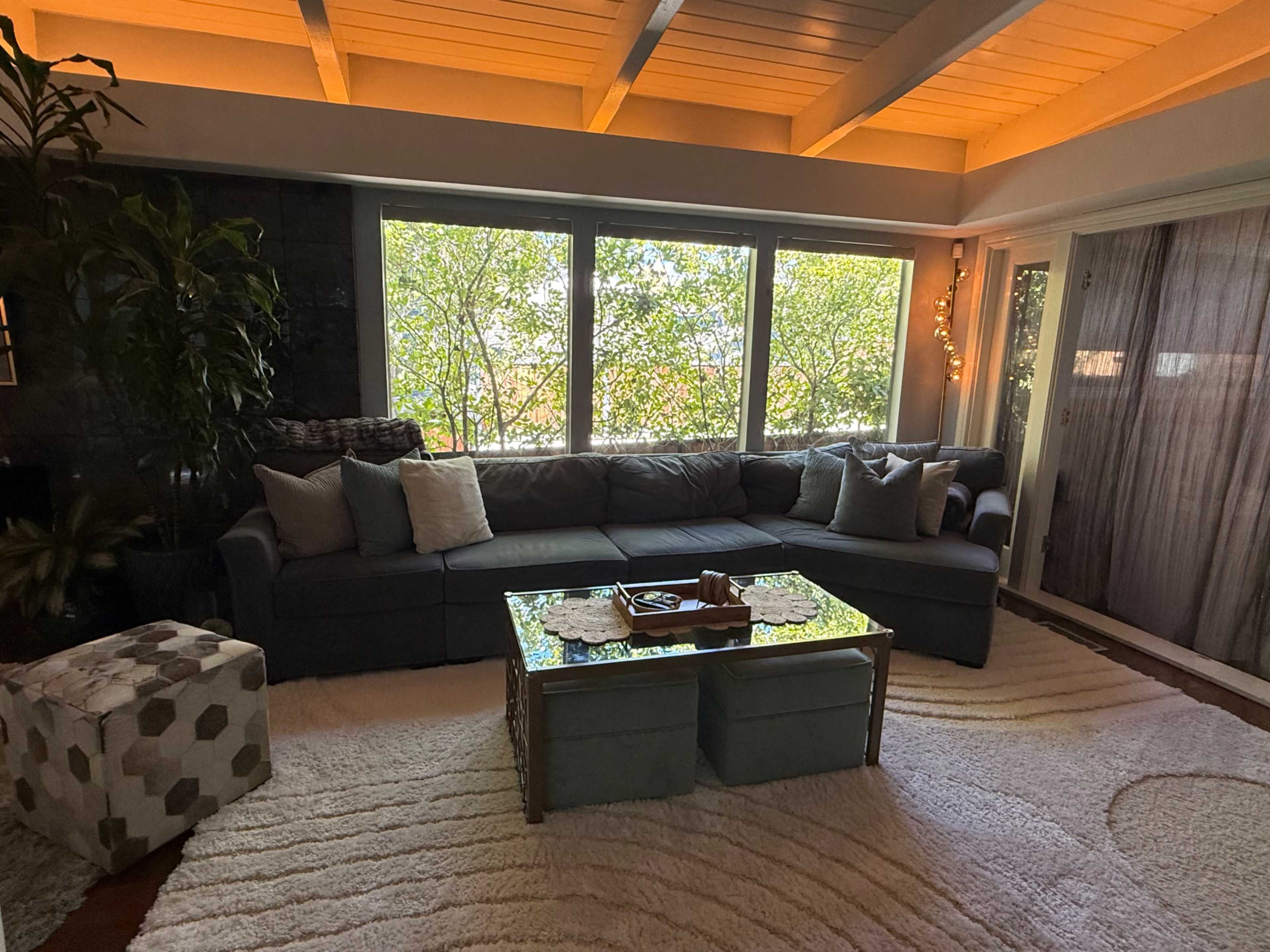 The image shows a modern living room with a large sectional sofa, a glass coffee table, and ample natural light coming through large windows, surrounded by greenery.