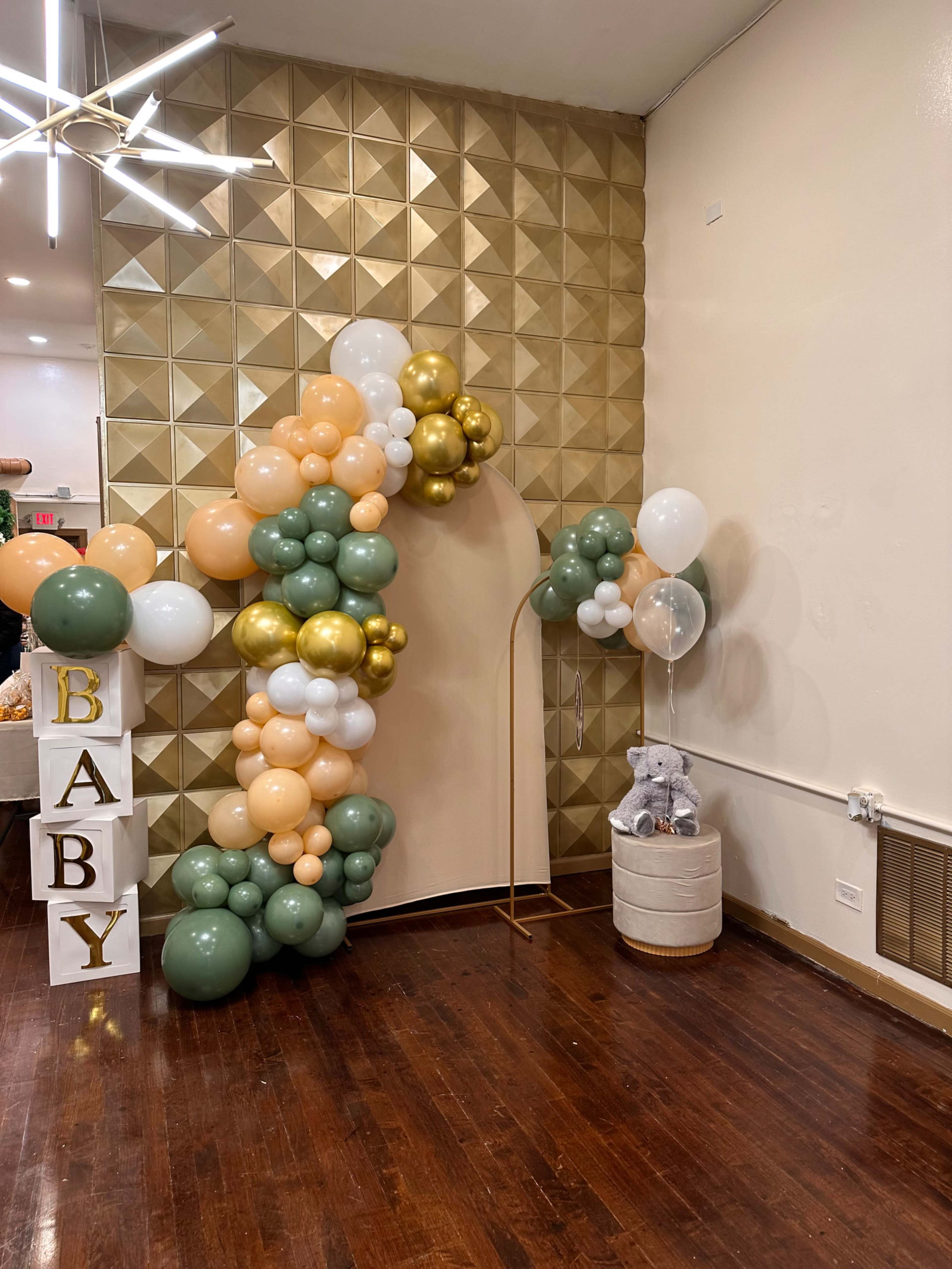 A decorative balloon arch of various colors next to a plush gray elephant and a white basket in a room with geometric wall panels.