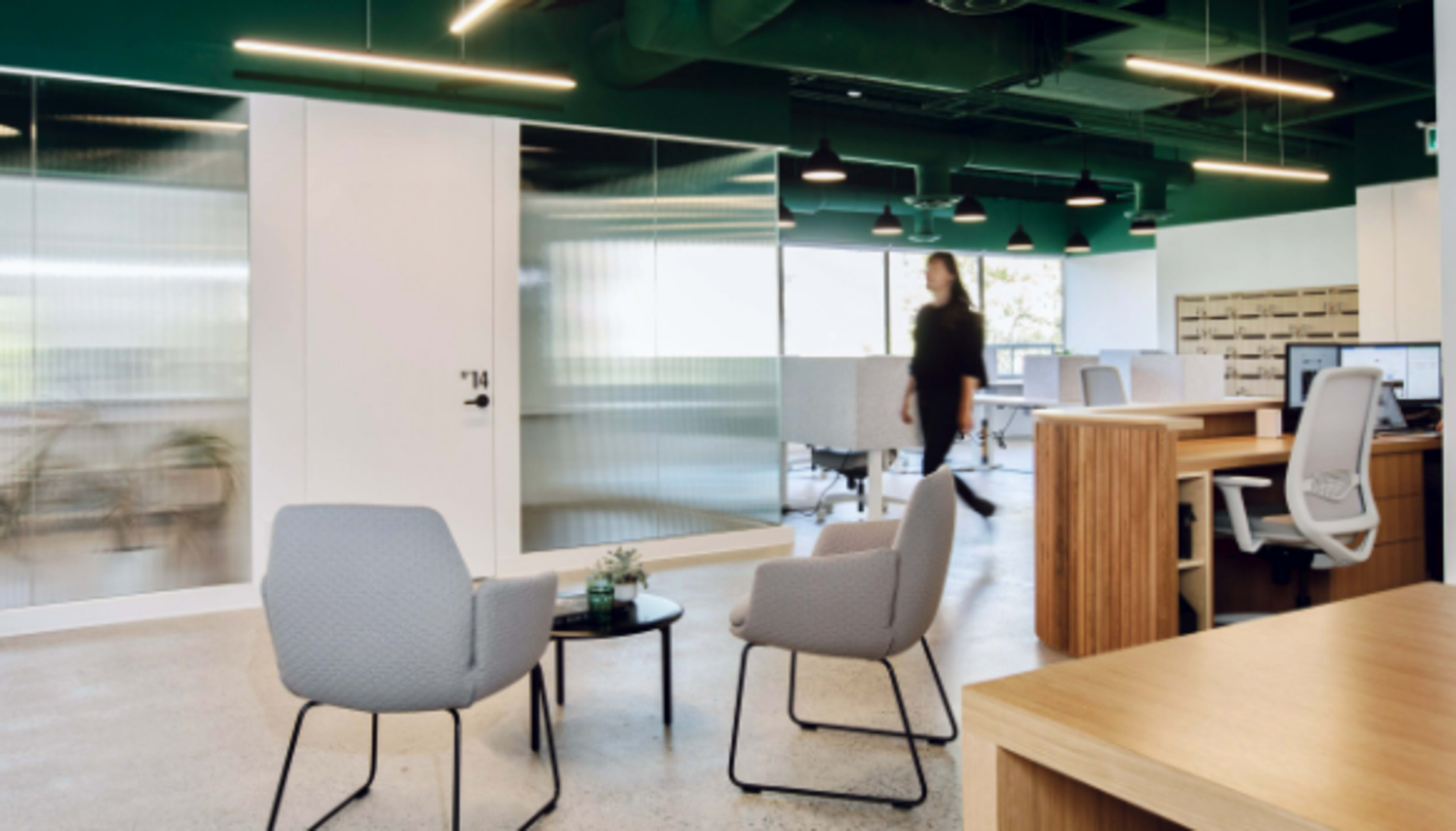 The image shows a modern office space with two gray chairs and a small table in the foreground, while a person walks past in the background amidst desks and glass partitions.