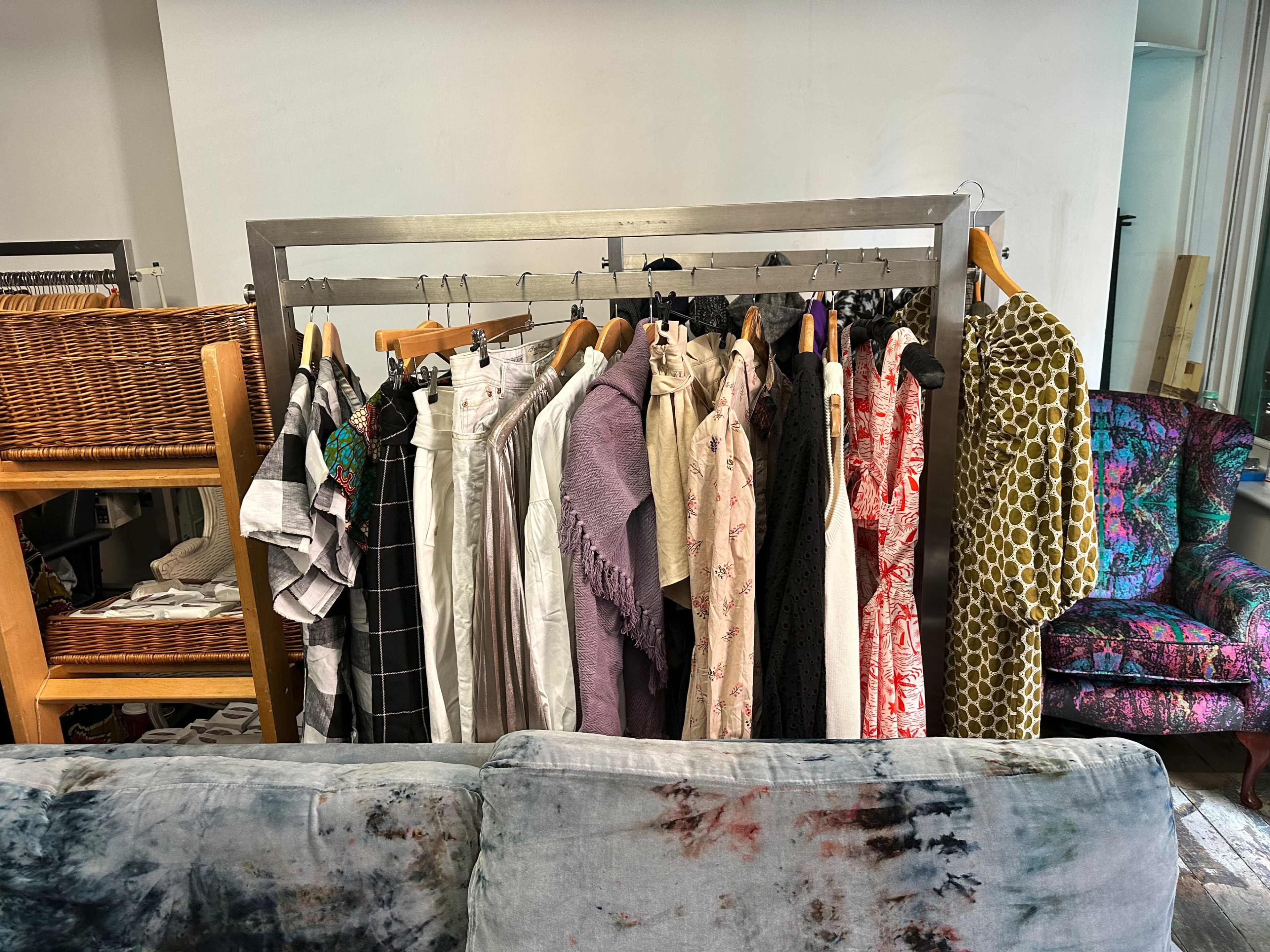 A clothing rack displays a variety of garments in a shop setting, with a couch in the foreground and woven storage nearby.