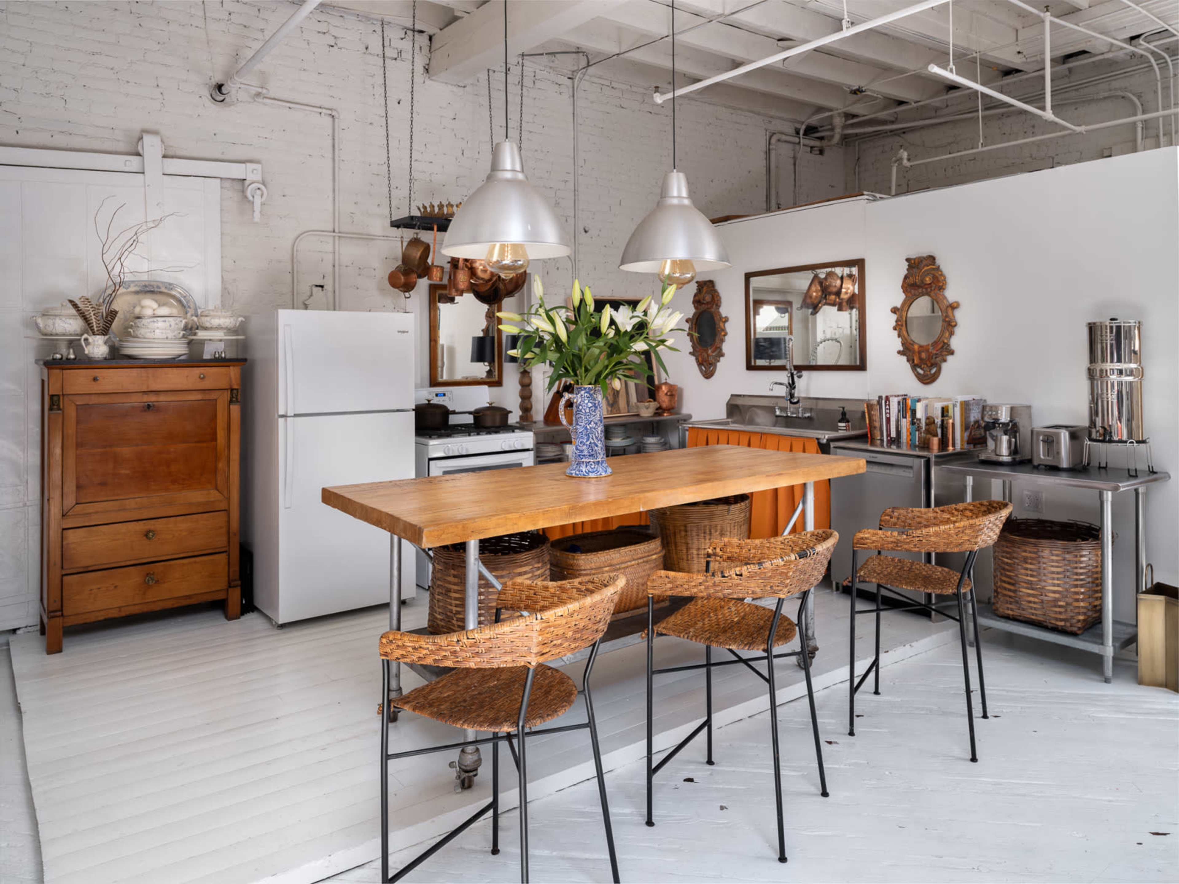 A modern kitchen with a wooden island, metal stools, a white refrigerator, and decorative wall mirrors, all set against a brick wall.
