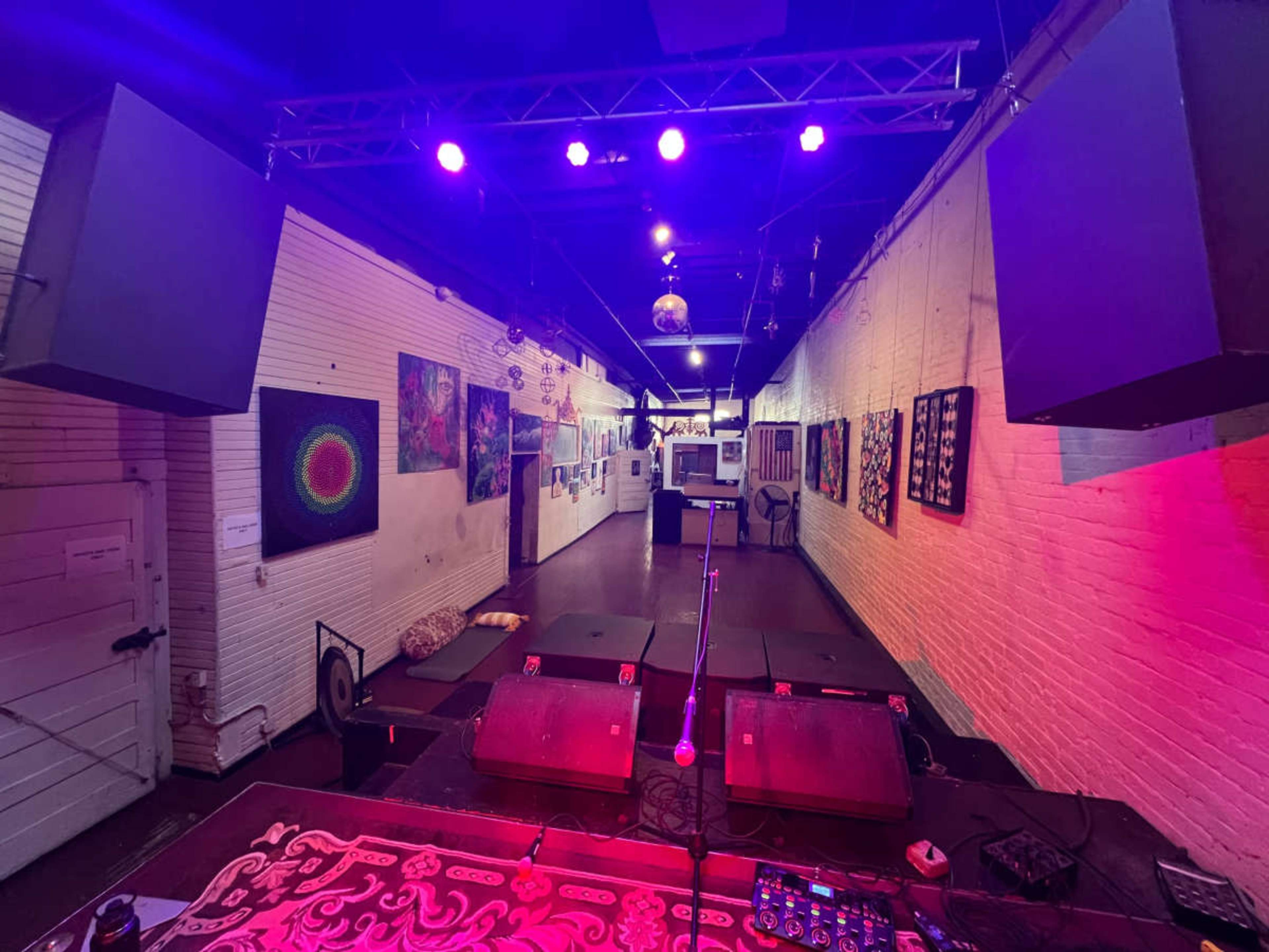 The image shows an empty performance space with colorful artwork lining the walls and a stage set up with speakers and a microphone.