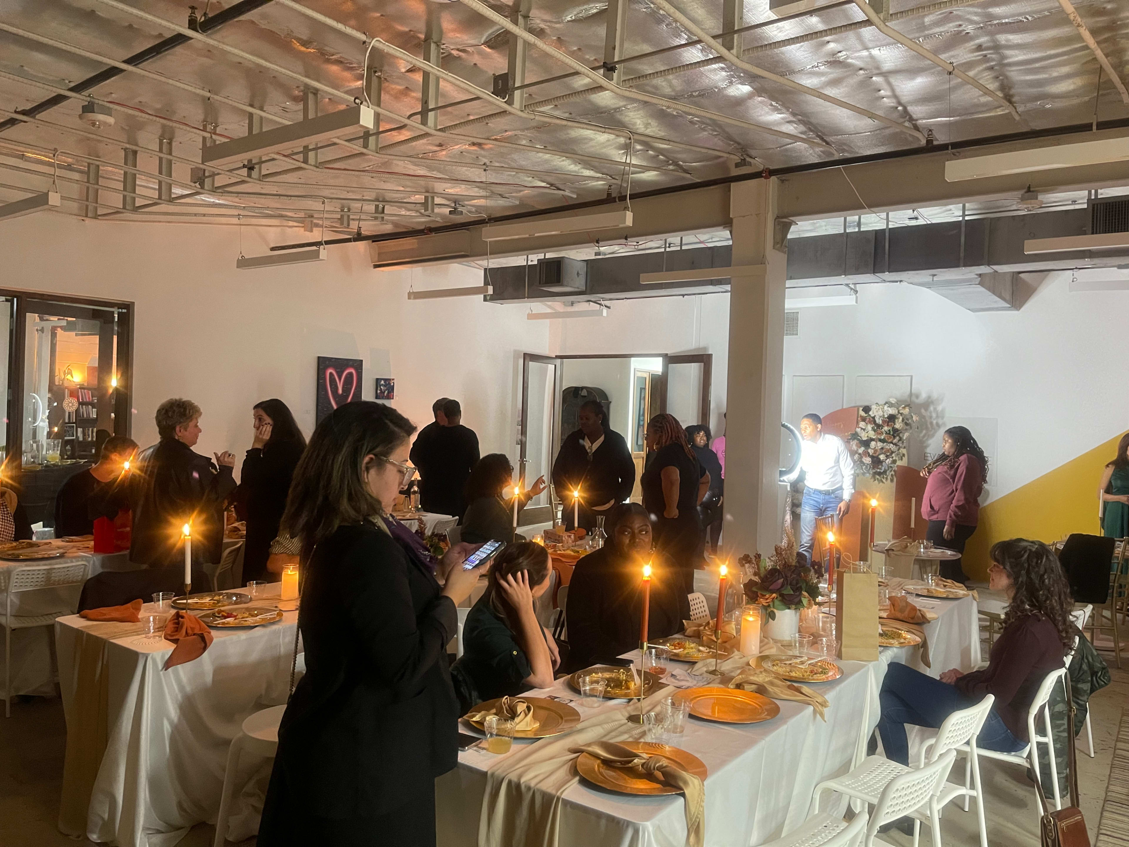 A gathering of people is seated and standing around elegantly set dining tables in a well-lit, decorated room.