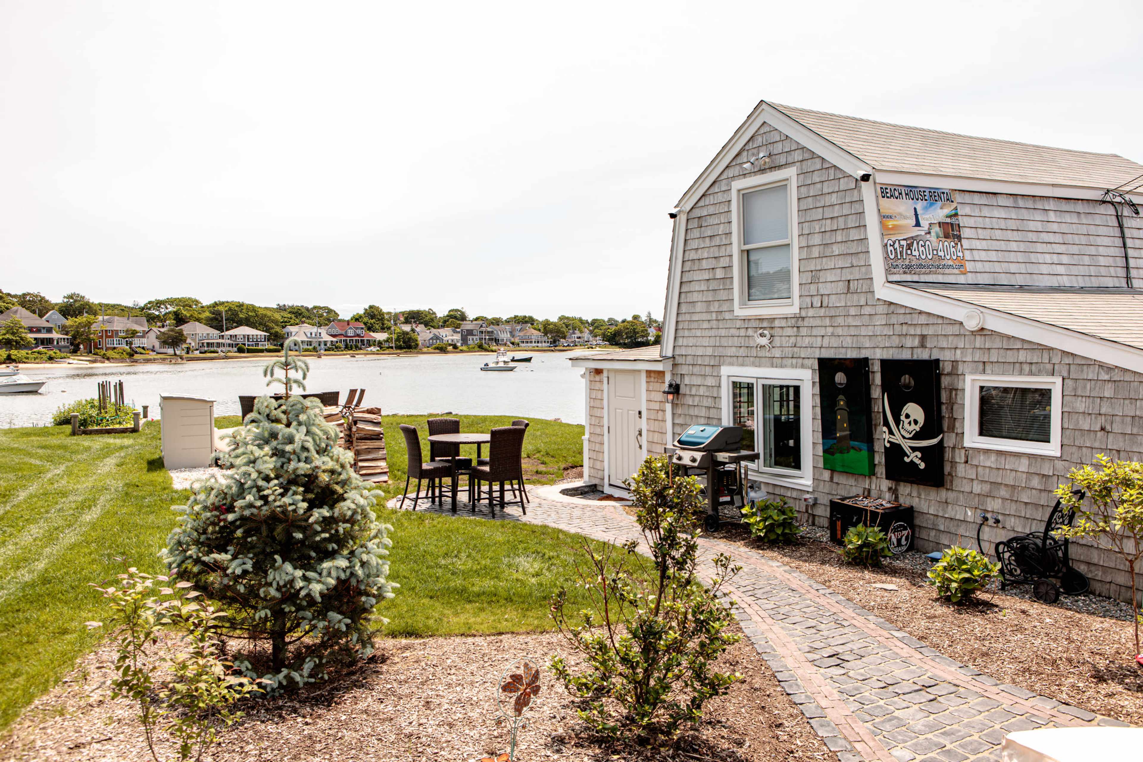 A waterfront house with a shingle exterior, a pirate flag, and outdoor seating overlooks a calm bay with boats nearby.