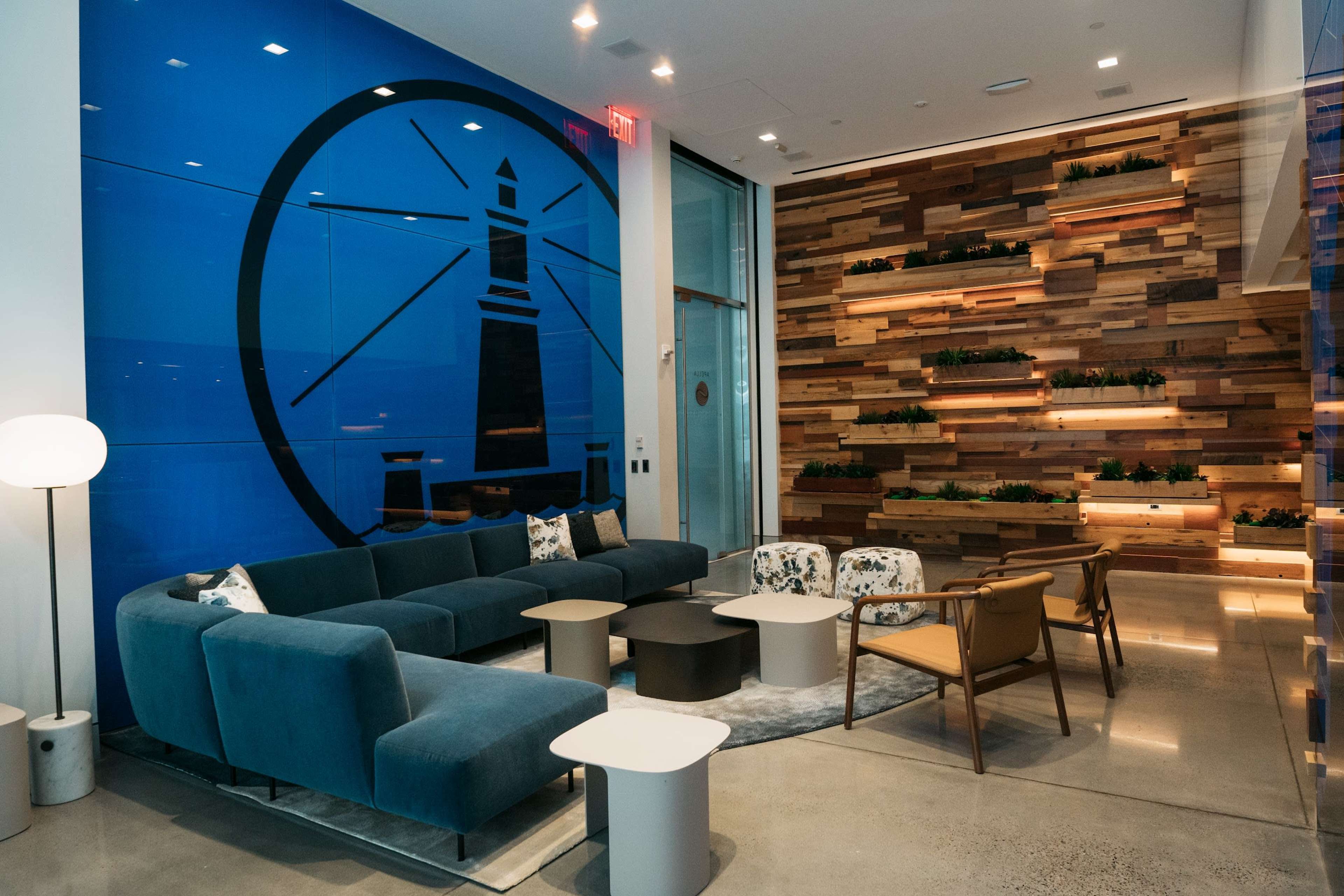 The image shows a modern lounge area featuring a blue accent wall with a lighthouse design, wooden planter shelves, and comfortable seating arrangements.