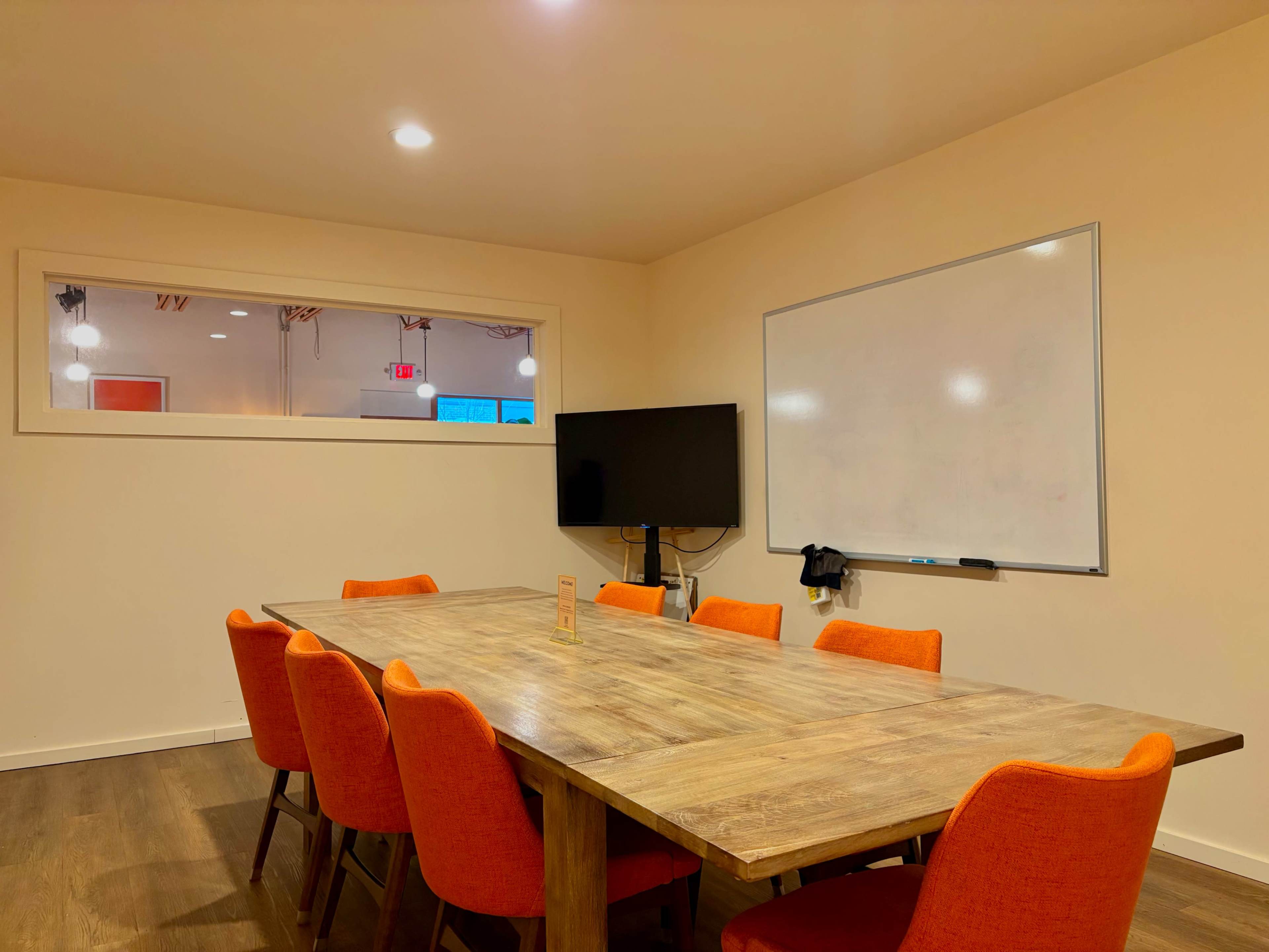 A large wooden table with eight orange chairs surrounds it in a meeting room, featuring a whiteboard and a television on the wall.