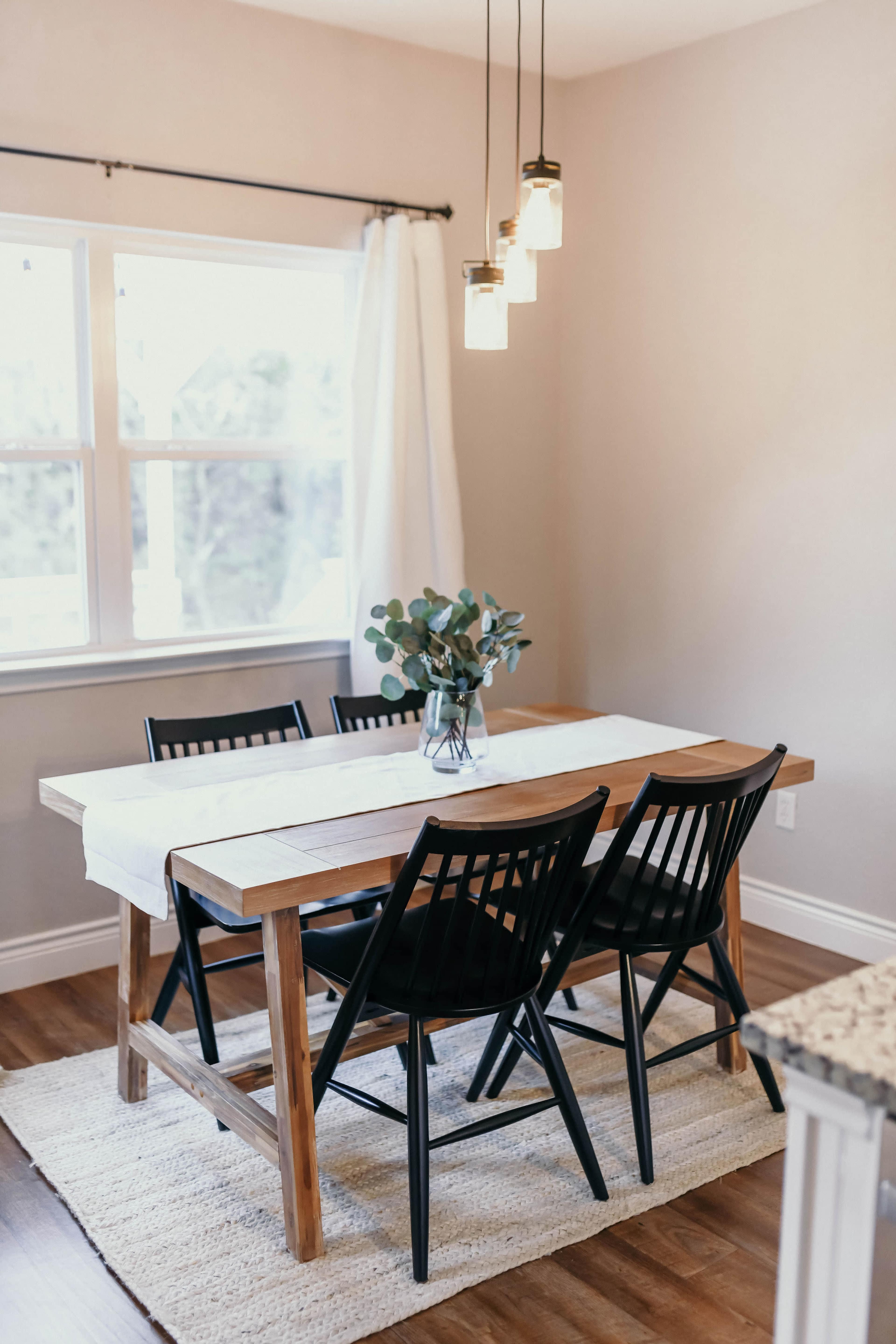 A dining room features a wooden table surrounded by four black chairs, with a vase of greenery at the center, illuminated by pendant lights above.