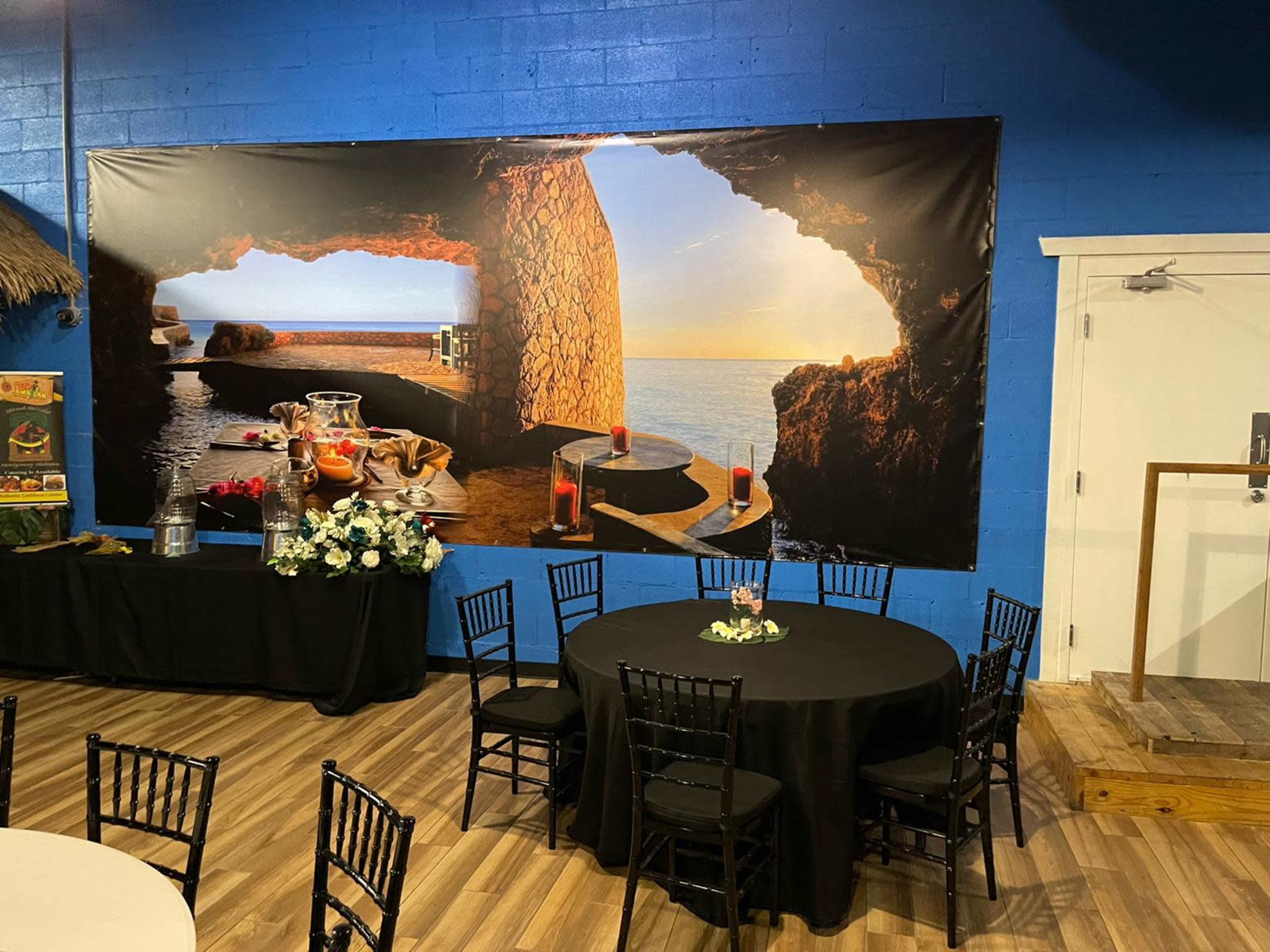 A large mural of a coastal cave scene on a blue wall, with black tables and chairs arranged in front of it.