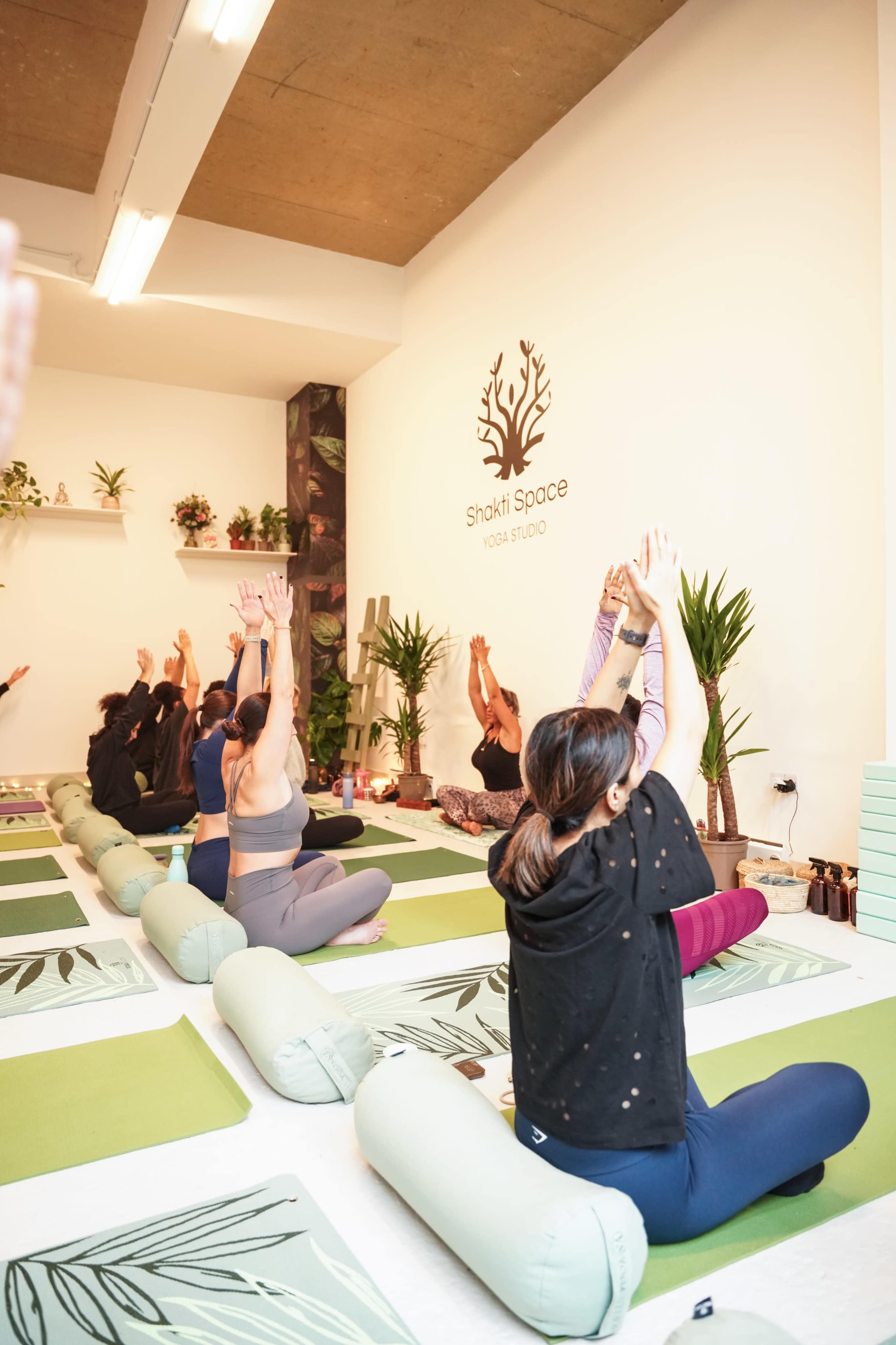A group of individuals is practicing yoga in a studio, with participants engaged in a seated pose while raising their arms.