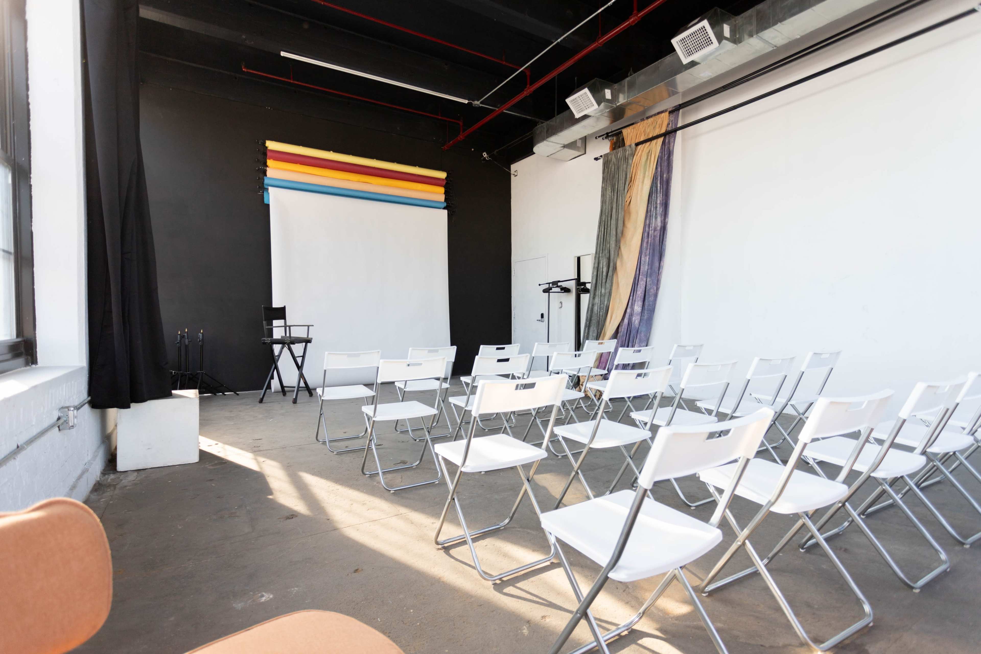 A minimalist room set up for an event, featuring rows of white folding chairs facing a projector screen against a black wall.